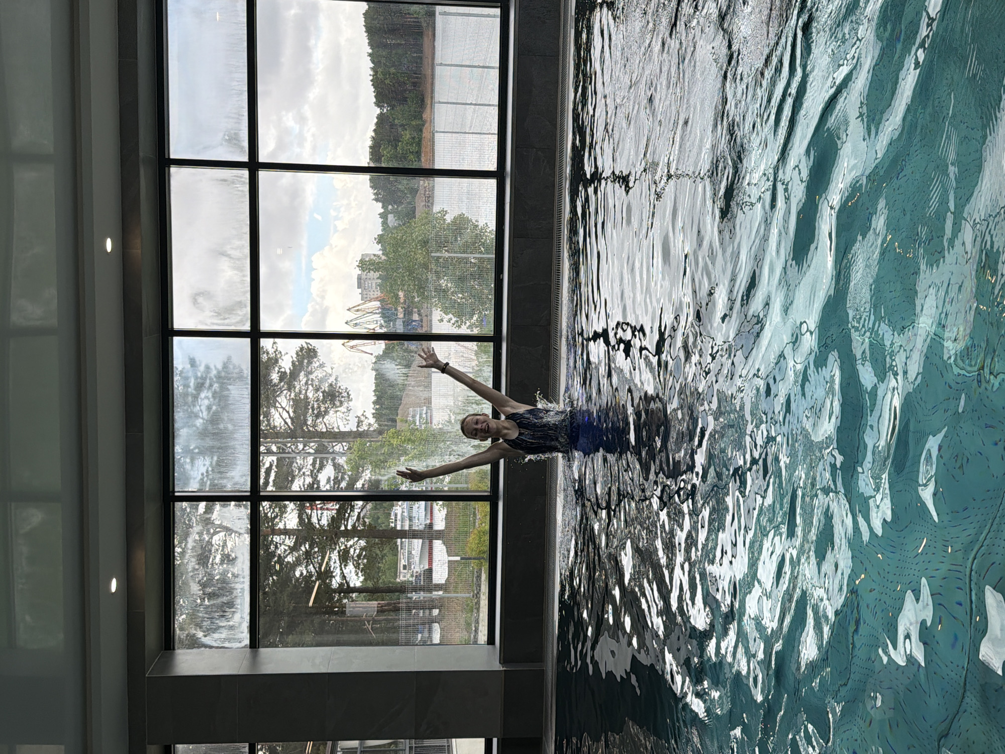 The image shows a person swimming in an indoor pool next to a large window. The person is in the center of the frame, with their arms outstretched and a smiling expression. The pool water is rippled and reflects light. Outside the window, there is a view of an amusement park or a construction crane, trees, and a body of water. Based on these landmarks, the location is most likely the pool in the Edem Hotel in the city of Chaikovsky, Perm Krai, Russia. The weather is overcast.