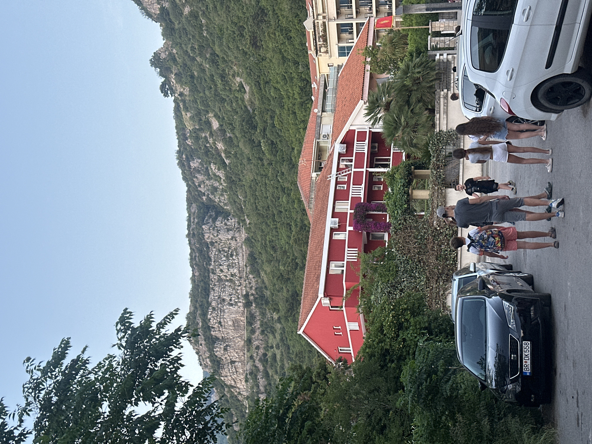 The image depicts a street scene in Budva, Montenegro, likely a residential area due to the presence of a red-painted building with white accents and a terracotta-tiled roof. The building is nestled amidst dense greenery and has a ladder visible against its side. A significant portion of the backdrop is dominated by a massive, rocky mountain, suggesting the town's proximity to a dramatic landscape.
On the street, a group of people are walking. There are at least six individuals visible, including two women with long hair, one wearing a white shirt and short white shorts, and the other a white shirt and short blue denim shorts. Several other individuals are visible, including a man and boy.
Parked on the side of the street are two cars: a white car and a black car. The black car has a license plate that reads "BR DK 558". The street itself appears to be narrow, indicative of a residential road. The scene is set during daylight, with a clear sky overhead.