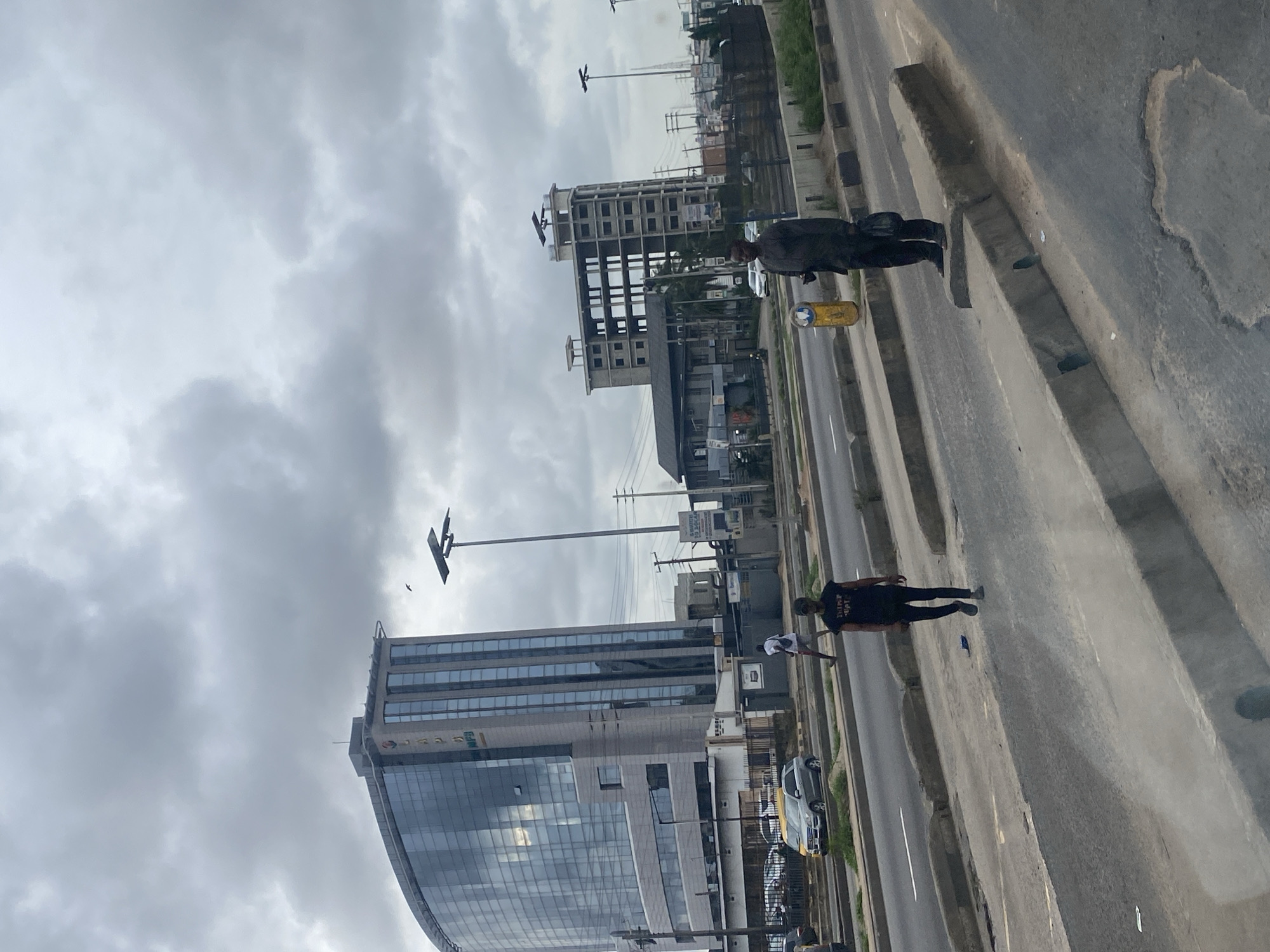 In Ikeja, Nigeria, under a cloudy sky, a wide asphalt road is visible. A curved, modern office building with a glass facade dominates the left side of the frame. To the right, an unfinished, multi-story building with exposed concrete floors and windows stands. Several vehicles, including a silver SUV and a yellow taxi, are on the road. Two men are standing on a concrete median in the foreground. One man, wearing a black t-shirt and dark pants, is walking away from the camera. The other man, in a dark outfit and holding a bag, is standing still, looking towards the right. A yellow cylindrical object is behind him. Streetlights and electrical poles are scattered throughout the background.