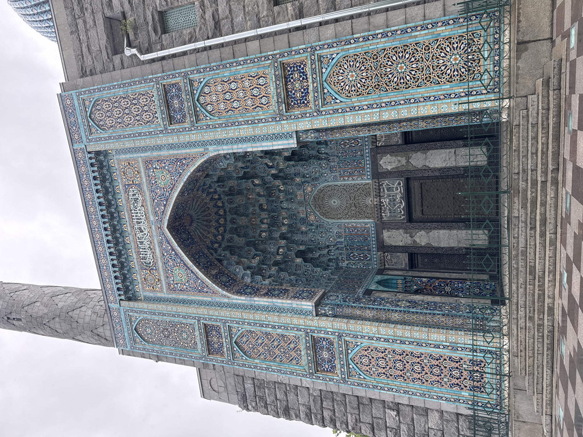 The image depicts the exterior of the Saint Petersburg Mosque, located in Saint Petersburg, Russia. The architectural style is reminiscent of Samarkand. The façade is adorned with elaborate mosaic tiles in shades of blue, turquoise, and beige, exhibiting intricate geometric and floral patterns. The main entrance is arched with the ceiling decorated with rows of muqarnas. Calligraphy in Arabic script is visible on the archway. The building is constructed with grey stone and brick. A minaret is partially visible on the left side. A fence runs along the entrance. Steps lead up to the entrance. The sky overhead is overcast.