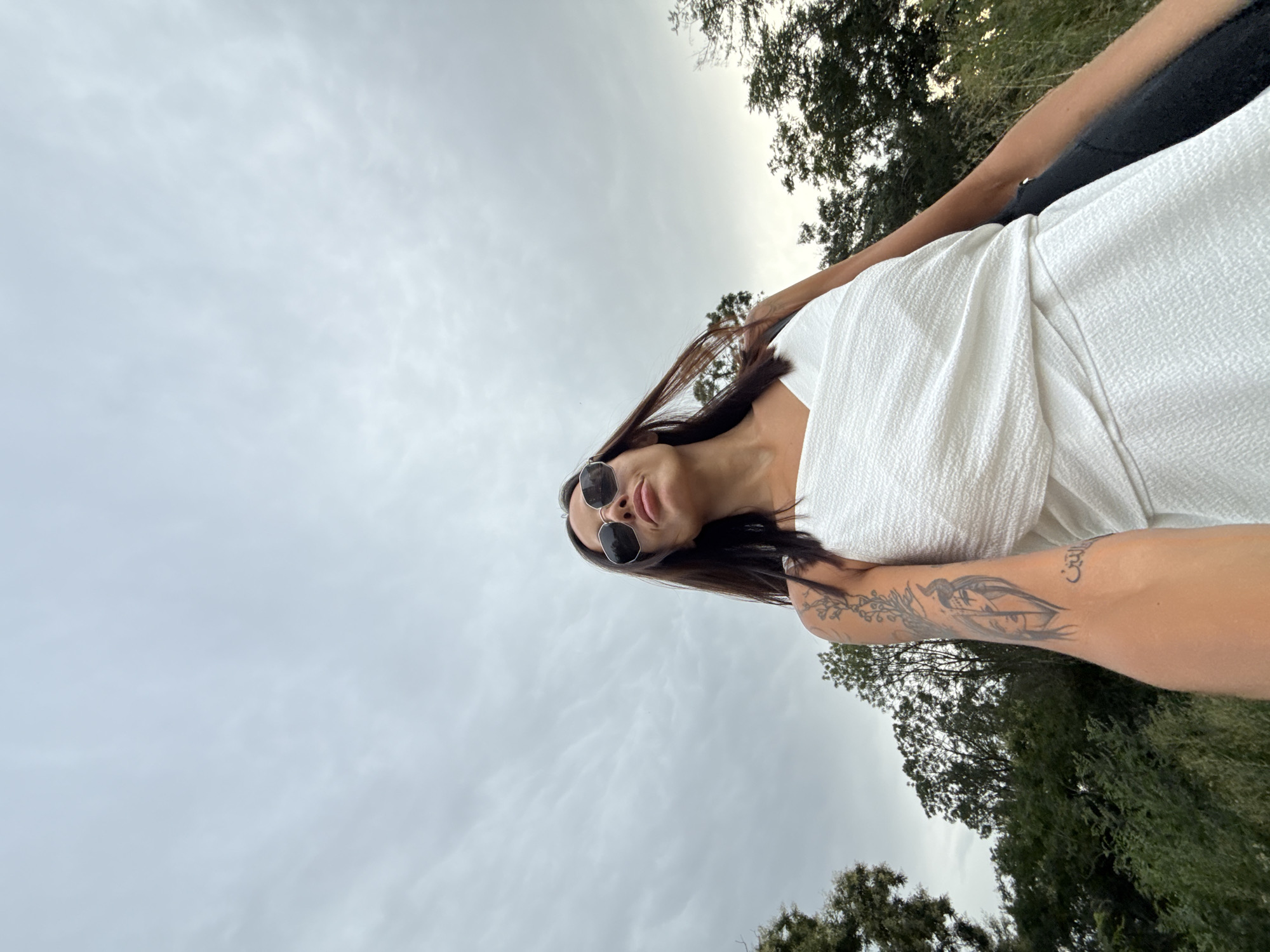 The image shows a woman in a white top and sunglasses, looking up at the sky. She has a tattoo on her arm. The background is a cloudy sky and trees. The location is the city of Pleven, Bulgaria, specifically the area around the Pleven Renaissance Window.