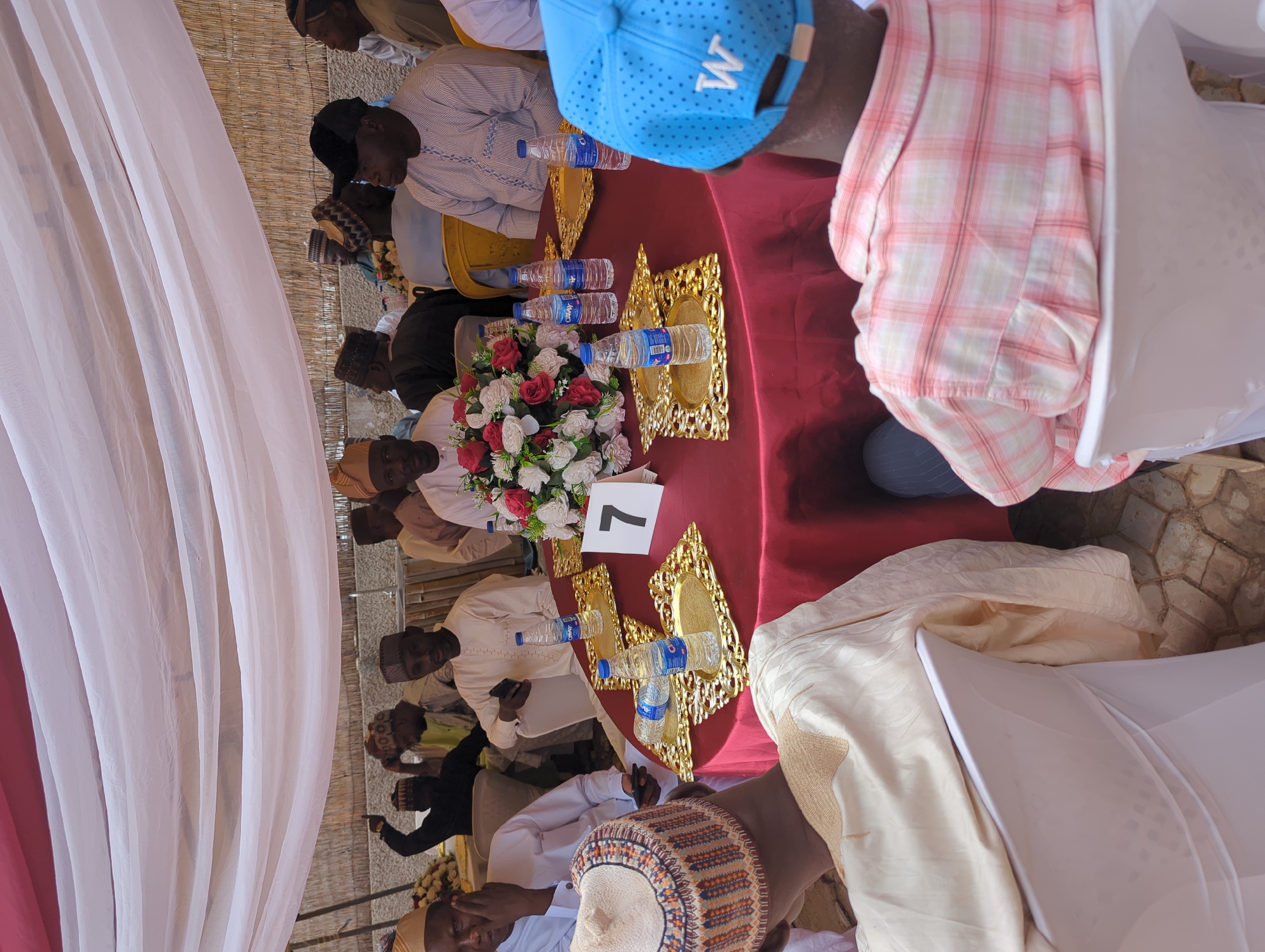 This image depicts a gathering of approximately 10-15 men under a temporary canopy structure, taking place in Kabala, Nigeria. The scene centers around a round table covered with a dark red tablecloth, equipped with a floral centerpiece of red and white roses.
**People:**
In the immediate foreground, two men are seated with their backs partially facing the camera. The man on the left wears a light-colored traditional garment with a patterned cap featuring brown, orange, and white hues. His right arm rests on a white chair cover. The man on the right wears a light blue baseball cap with a white 'W' logo on the back and a pink and white plaid shirt; his left shoulder is visible, also resting on a white chair cover.
Around the round table, multiple men are seated. A man directly opposite the viewer wears a light-colored traditional outfit and matching cap, looking forward. To his left (viewer's right), a man in a light-colored striped shirt and a black baseball cap is seated, facing left. To his right (viewer's left), another man in a dark traditional outfit and cap is partially obscured, with his hand near his face. Further back and to the left, a man is visible with his right arm raised and index finger pointing upwards. The majority of individuals are dressed in traditional Nigerian attire, including long robes and various caps, while some wear Western-style clothing. Several men have mobile phones in their hands or placed nearby.
**Objects and Setting:**
The red-covered table features multiple ornate, gold-colored rectangular trays, each holding a clear plastic bottle of "CWAY" brand water. A white tent card displaying the number "7" in black is positioned on the table to the right of the floral centerpiece. The chairs are covered with white fabric. The gathering is situated under a white fabric canopy, draped in gentle folds across the upper portion of the frame, providing shade. In the background, behind the seated individuals, a light-colored wall or partition made of woven, natural material (possibly bamboo or reeds) is visible. The ground beneath the chairs is paved with irregular, light-toned stones or tiles. The scene is illuminated by natural daylight.