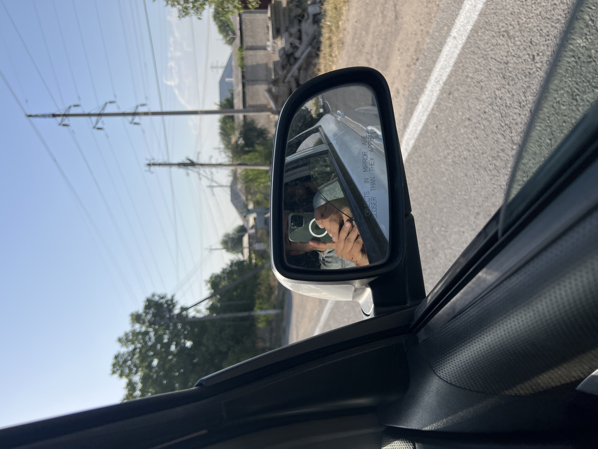 The image captures a view from inside a vehicle traveling on a paved road, as seen through the driver's side window and side-view mirror. The side-view mirror reflects a person holding a smartphone, taking a selfie. The phone's screen is visible, showing the captured image, and the person's hand with a ring is also visible. The mirror itself has the warning text "OBJECTS IN MIRROR ARE CLOSER THAN THEY APPEAR" inscribed on its lower edge.
The exterior environment visible through the window includes a road with a white line marking the lane, a shoulder of gravel and dry vegetation, and a row of trees with green foliage. In the background, several structures, likely houses, are partially visible, along with power lines supported by tall metal towers. The sky is clear and blue. The context provided suggests this scene is located in the city of Yangikurgan, Uzbekistan.