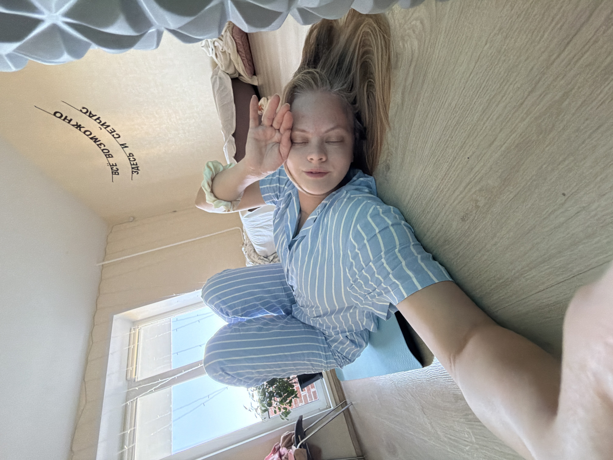 A young woman with long blonde hair, wearing blue and white striped pajamas, is sitting cross-legged on a light brown wooden floor in a room with a window. She has her eyes closed and her left hand raised to her face. In the background, a window with a view of buildings and sky is visible. On the ceiling, there is text in Russian that reads "Здесь и сейчас всё возможно." The room appears to be in Izhevsk, Russia.