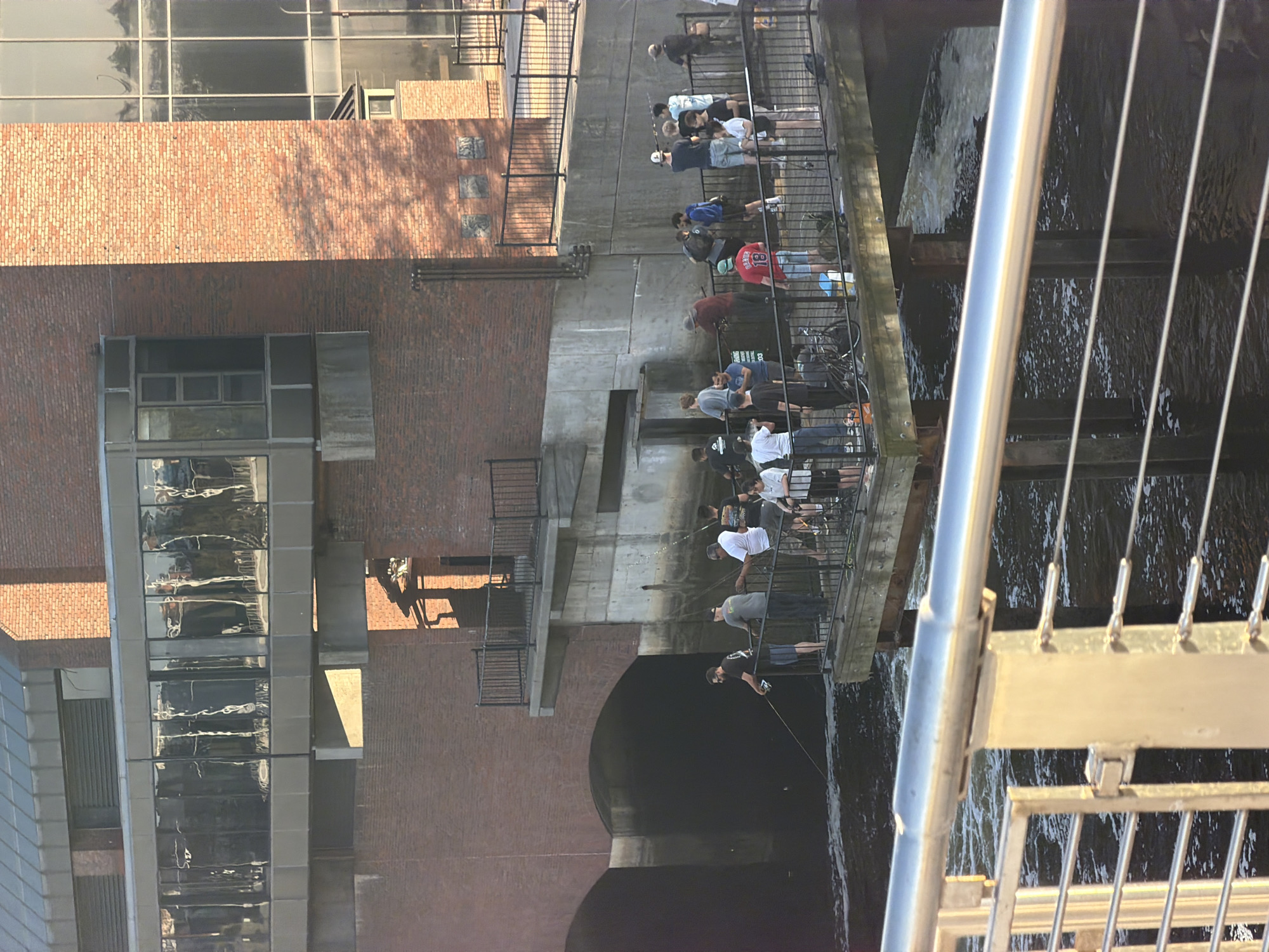 The image shows a scene in Boston, Massachusetts, United States. Several people are standing along a walkway and fishing in the water below. The walkway is bordered by a metal railing. The Charles River flows beneath the walkway. A brick building and a modern glass structure are visible in the background.