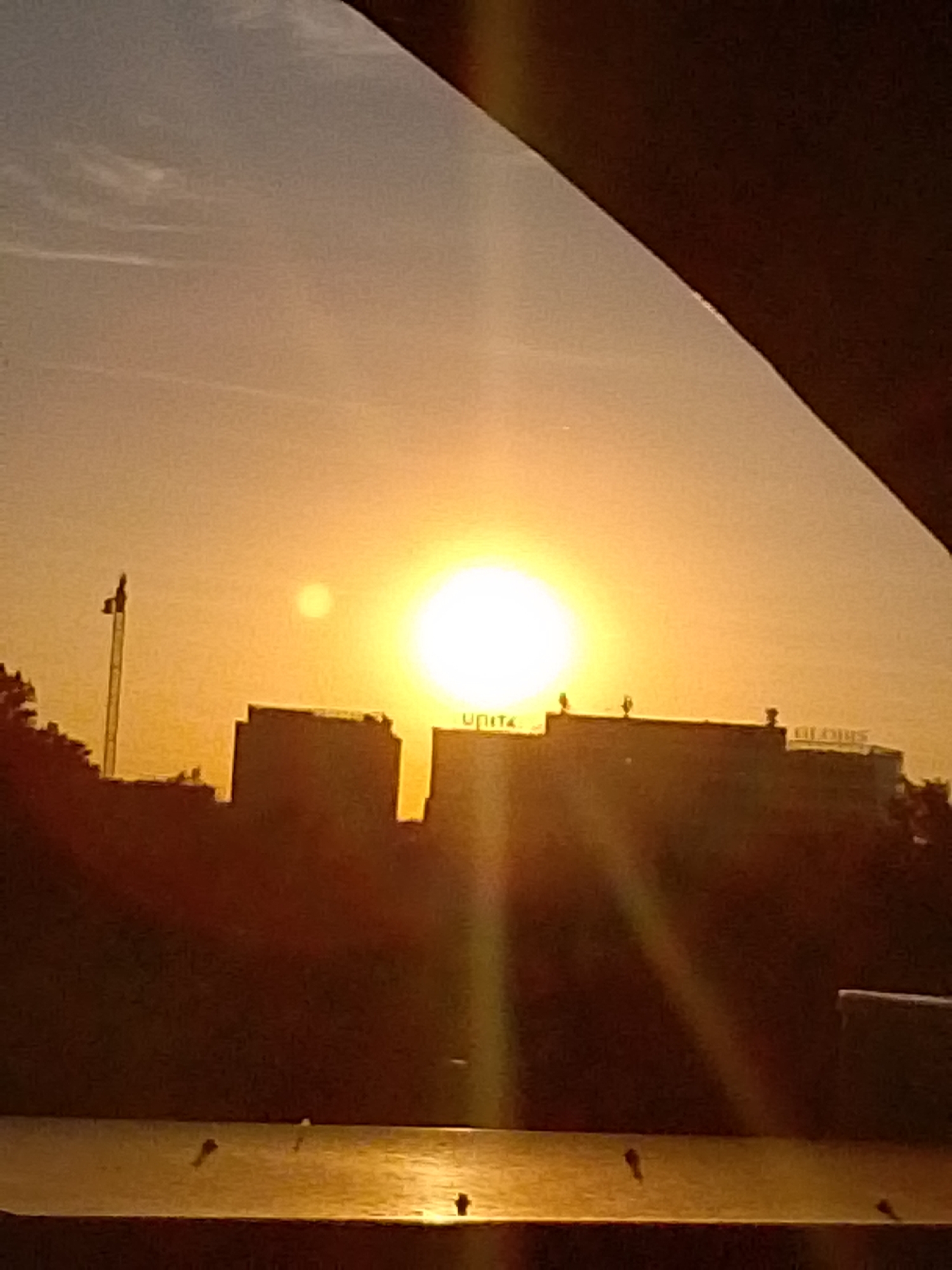 A sunset casts a warm, golden glow over an urban landscape. In the foreground, a dark, curved structure, possibly a building overhang or a vehicle roof, obscures the top of the frame. Below this, a body of water, likely a river or canal, reflects the warm light, with several dark, indistinct objects visible on its surface, possibly small boats or debris.
Dominating the mid-ground are silhouetted buildings, their shapes softened by the low light. The largest building on the right features illuminated lettering that appears to spell "GLOBUS". Another building to the left has partial lettering, possibly "UNITU". A tall, slender pole with a dark object at its apex, resembling a flag or antenna, stands to the left of these structures. The sky is a gradient of pale orange and yellow, with subtle wisps of clouds. The sun itself is a bright, luminous orb, slightly off-center to the right, with a smaller, fainter light source visible to its left, possibly a reflection or another celestial body.
The scene is set in Wrocław, Poland, with the buildings likely being part of the city's skyline. The overall impression is one of a serene, late-day urban panorama.