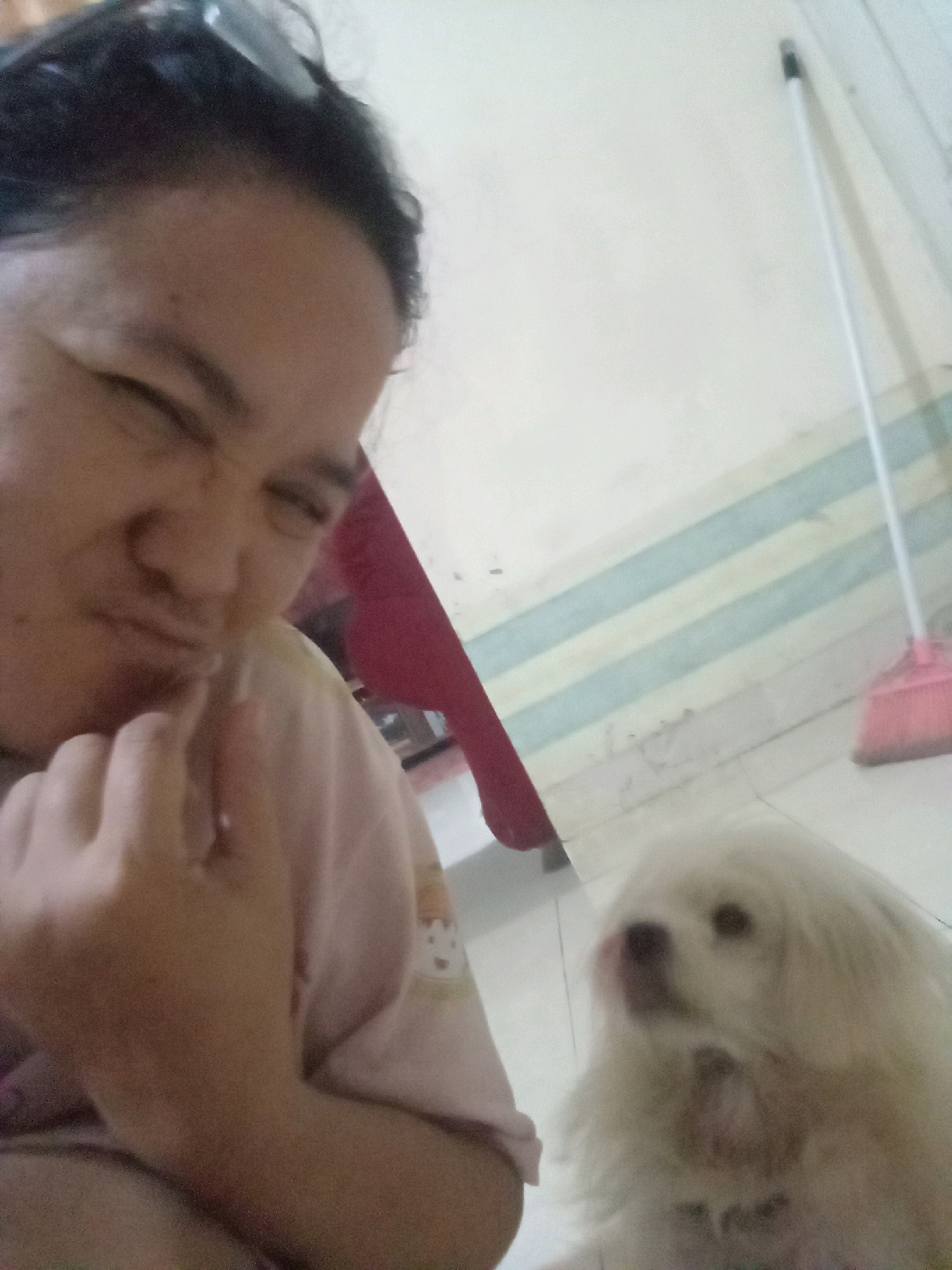 A person is taking a selfie with a white dog indoors. The person is in the foreground on the left, with their left eye closed and their right eye looking at the camera. They are wearing a light pink t-shirt with a graphic on it. Their left hand is raised, with their index finger touching their chin. The dog is in the right foreground, also looking towards the camera. It is a small white dog with long fur. In the background, a pink broom with a white handle is leaning against a wall. The wall has horizontal blue and white stripes. The floor appears to be tiled. The scene is lit by indoor lighting. The context of this image being taken in Jambi, Indonesia, is not evident from the visual information alone.