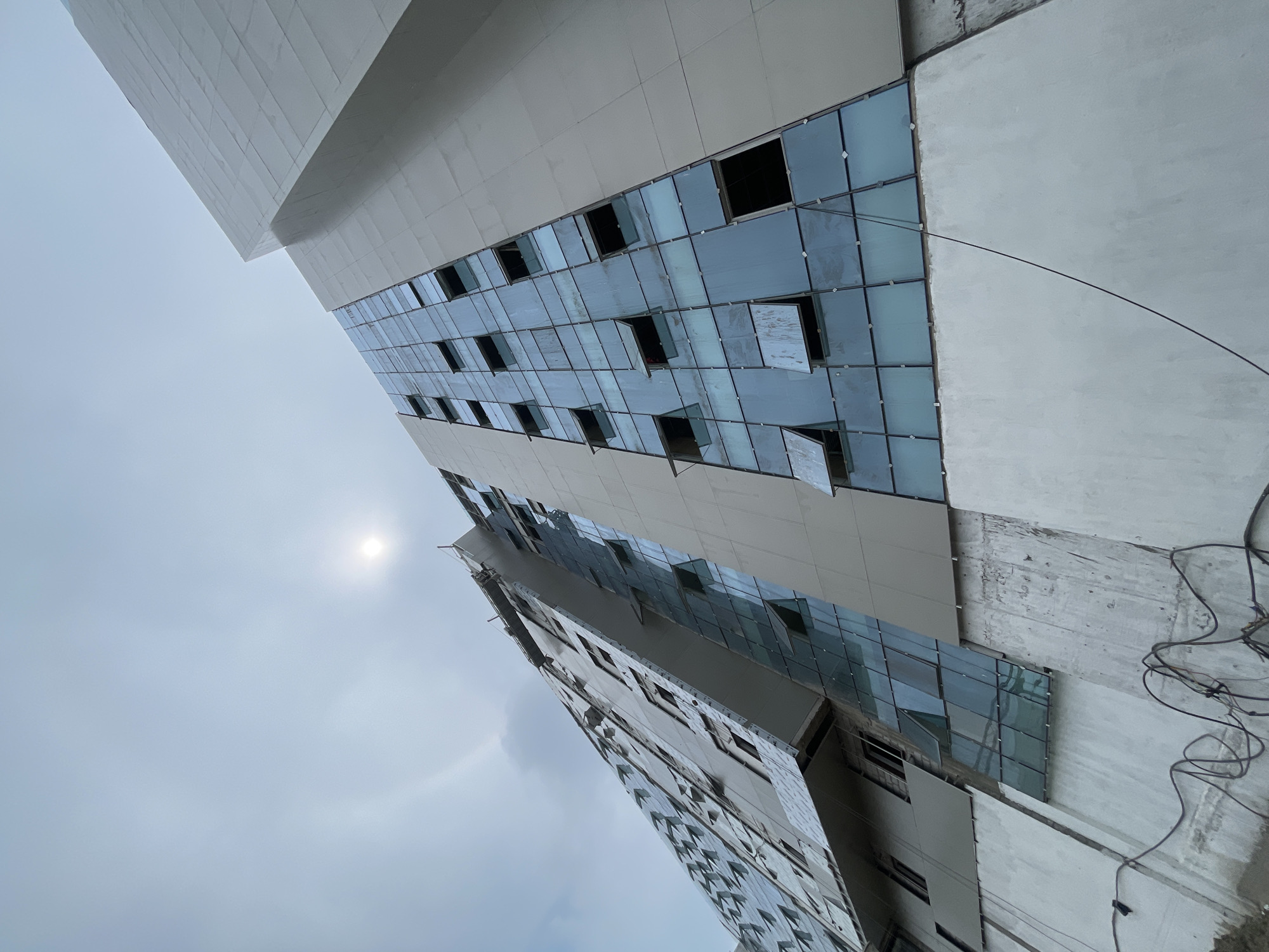 The image shows a tall, modern building under construction in New Delhi, India. The building has a facade composed of large, reflective glass panels and some sections covered in light-colored panels. Several of the glass panels are open windows, suggesting the building is either nearing completion or undergoing some form of inspection or maintenance. The sky above is overcast with a bright sun visible, indicating daytime. At the base of the building, unfinished concrete walls are visible, along with some electrical wiring, further confirming the construction status. The perspective is from a low angle, looking upwards at the imposing structure.