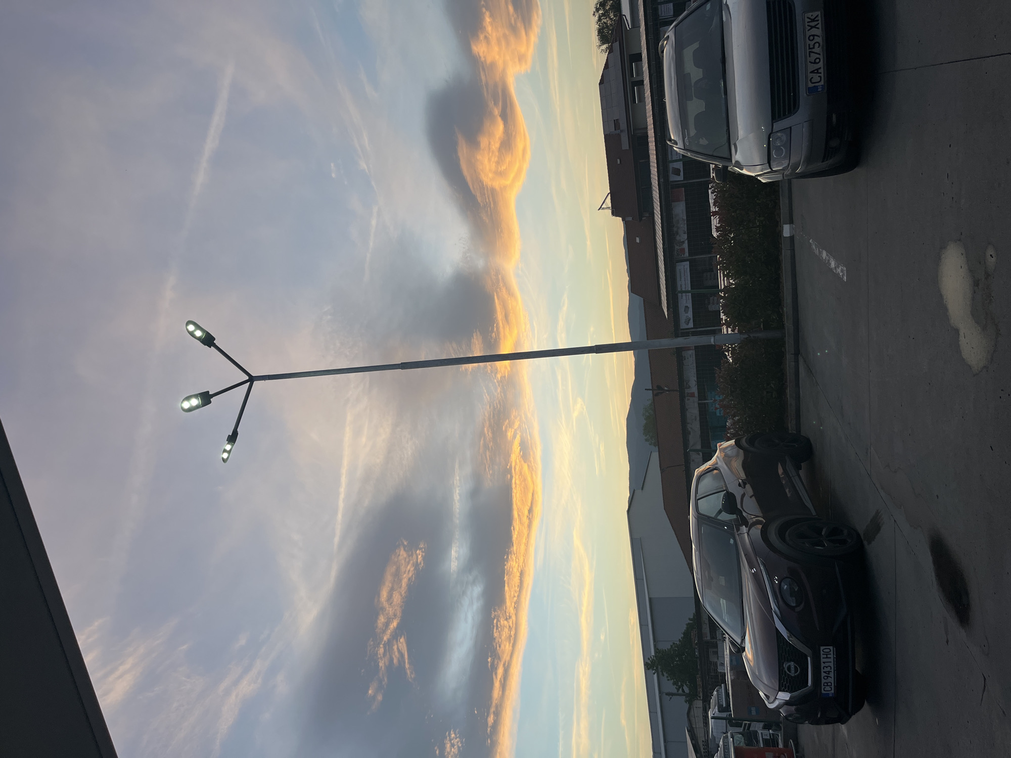 The image shows a parking lot with two cars, a silver van with the license plate "CA 6759 XK" and a burgundy Nissan Juke with the license plate "CB 9431 HO", in Gotse Delchev, Bulgaria. A street light stands prominently in the middle. The sky is filled with clouds illuminated by the setting sun. Buildings are visible in the background behind the cars.