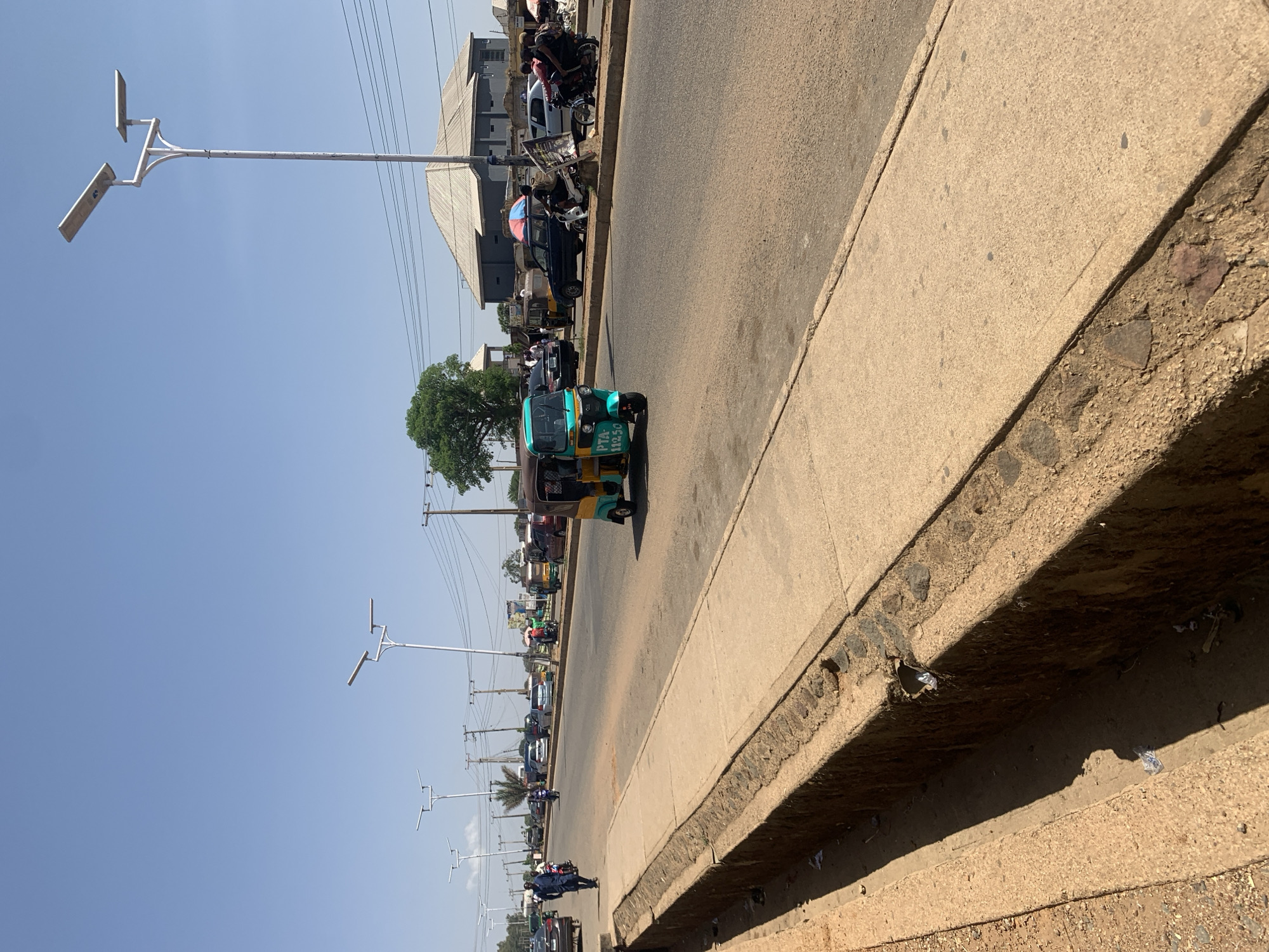 The image captures a street scene in Rafin Par, Nigeria. A three-wheeled vehicle, painted in shades of green and yellow, is traveling on the right side of the road. It bears the markings "PTA - 11250". The street is lined with other vehicles, including cars and motorcycles. Pedestrians are visible on the sidewalks. A tree provides shade near the road. Streetlights are visible along the road. The sky is clear and blue. The sidewalk and roadside area appear weathered.