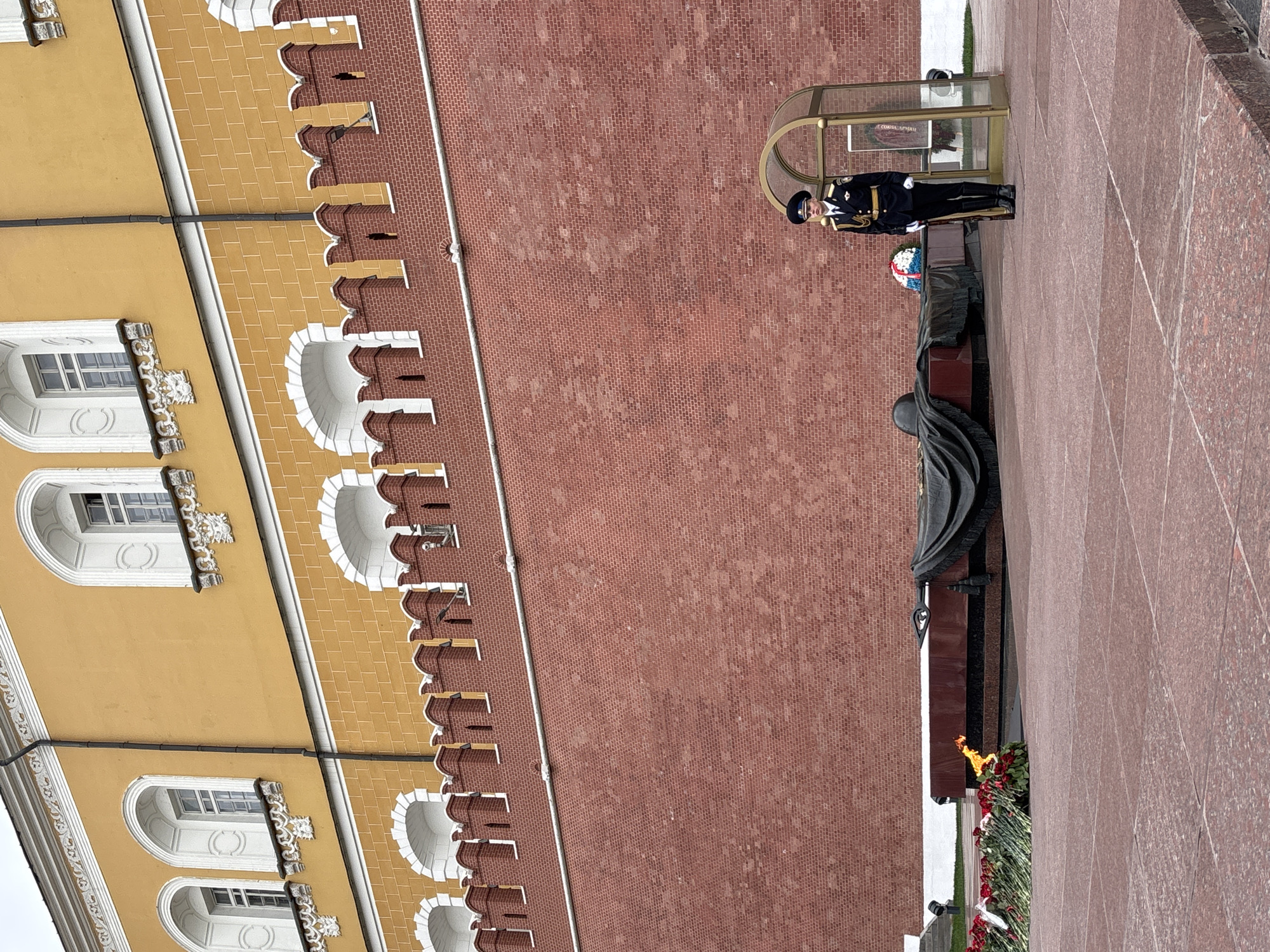 The image depicts the Tomb of the Unknown Soldier at the Kremlin Wall in Moscow, Russia. A guard stands at attention outside the glass booth. The eternal flame burns in front of the tomb, which is adorned with flowers. The yellow building is part of the Kremlin. The Kremlin wall is red brick. The pavement in the foreground is red granite.