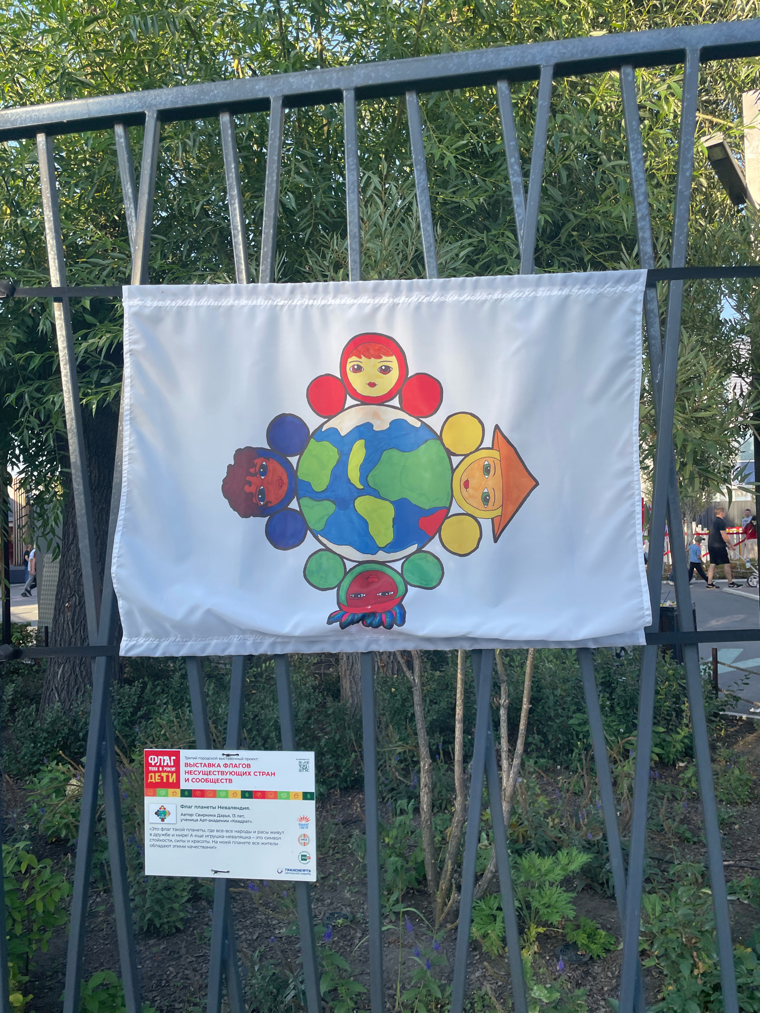 The image shows a white flag with a drawing of the Earth at its center, surrounded by five colorful figures resembling matryoshka dolls with different facial features and ethnic representations. The flag is attached to a metal fence. Below the flag, there is a sign with text in Russian. The sign is white with red and green accents, and it describes the flag as "Flag of the Planet Nevralandia," created by Daria Svirina, aged 13. The text explains that the flag represents a planet where all peoples and races live in friendship, symbolizing the matryoshka doll as an unbreakable bond and a symbol of these qualities. The background shows greenery and trees, suggesting an outdoor setting. In the blurred background, there are people walking, and a street scene is visible, indicating an urban environment. The presence of the sign with the description of the flag and the context of an exhibition of imaginary flags suggests this is likely part of a public art installation or event in the city of Omsk, Russia. The fence appears to be part of a park or public space.