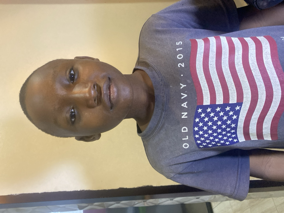 A young Black boy with close-cropped hair is depicted from the chest up. He has brown skin, and his face is turned slightly toward the viewer. He's wearing a gray t-shirt with a print of the American flag on the front. The words "OLD NAVY" and "2015" are also visible on the shirt. The background appears to be a light yellow or beige wall. The image is rotated counterclockwise, placing the wall at the left and the boy at an angle.