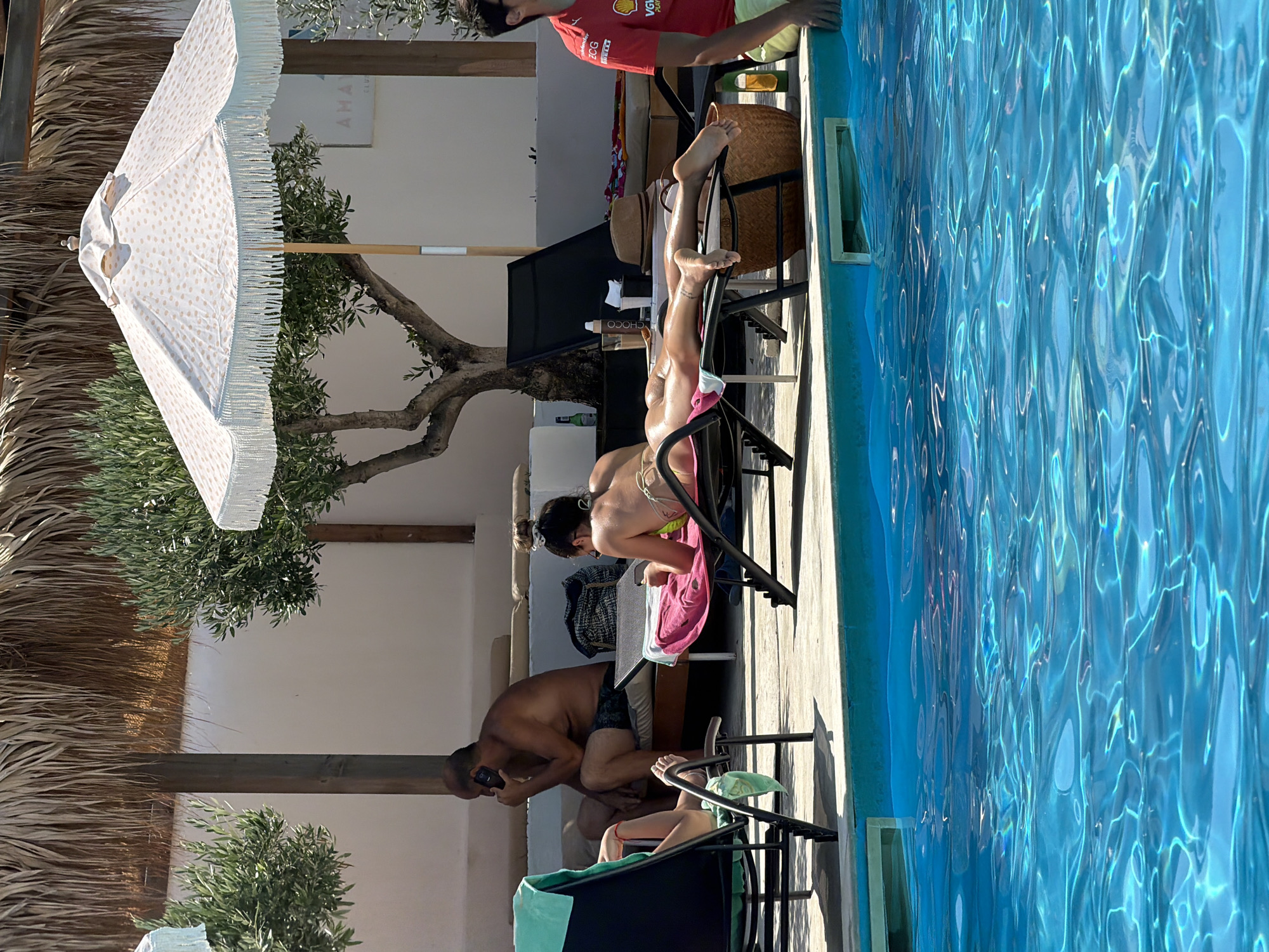 The image depicts a poolside scene in Burgas, Bulgaria, on a sunny day. In the foreground, a man in a red shirt with a logo is seated on the edge of the pool. Behind him, a woman is sunbathing on a lounge chair, wearing a bikini. Further back, another person is seated on a lounge chair and is on the phone. There are lounge chairs, umbrellas, and vegetation surrounding the pool. The water of the pool is a clear blue with ripples.