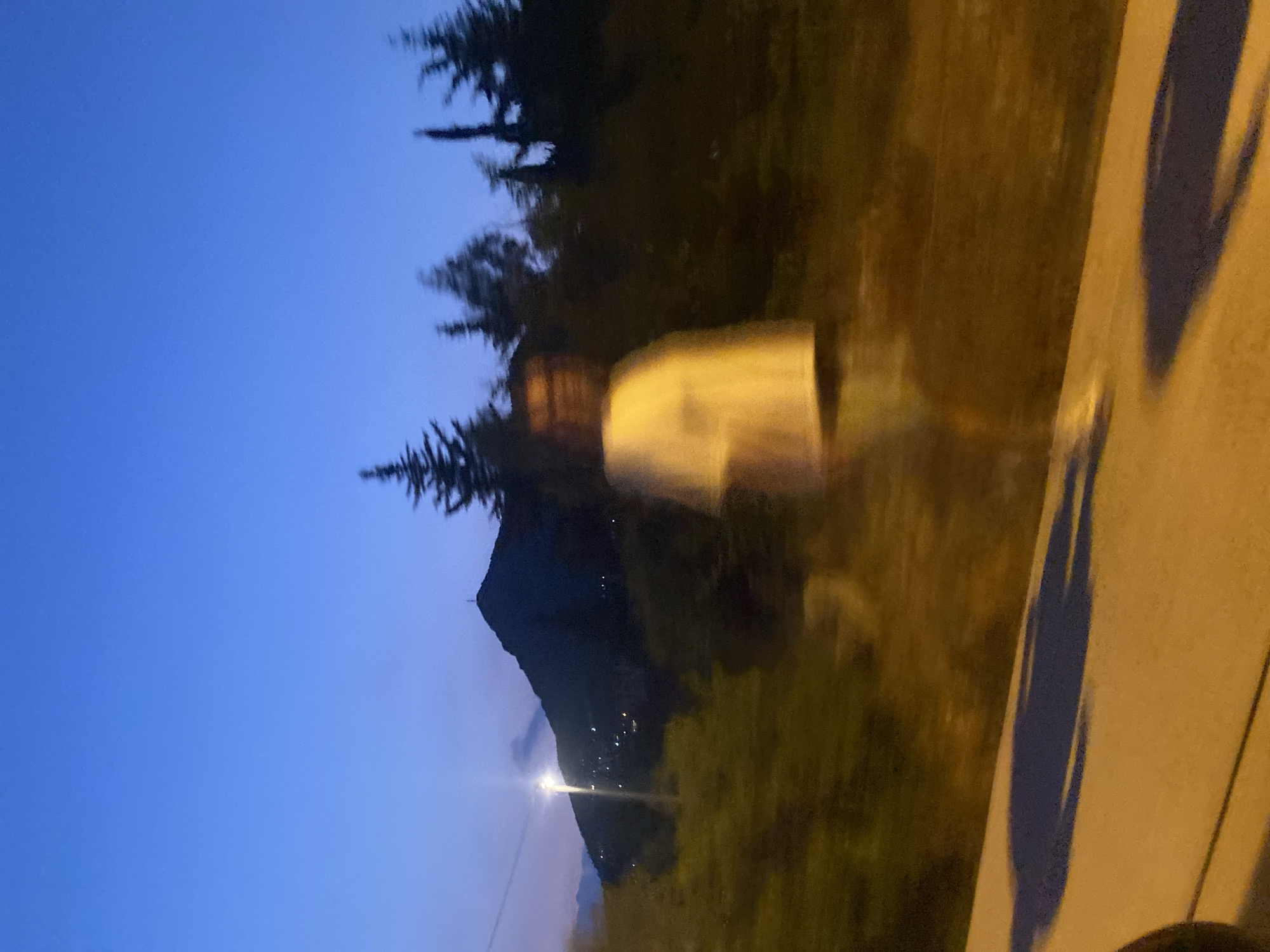The image captures a blurry silhouette of a person standing near a road at dusk. In the background, a mountain looms, illuminated by a streetlamp and dotted with distant lights, suggesting a settlement. Trees and foliage flank the road, with the sky a deep blue transitioning to the darker hues of twilight. The overall impression is one of movement and low light conditions, possibly captured from a moving vehicle.