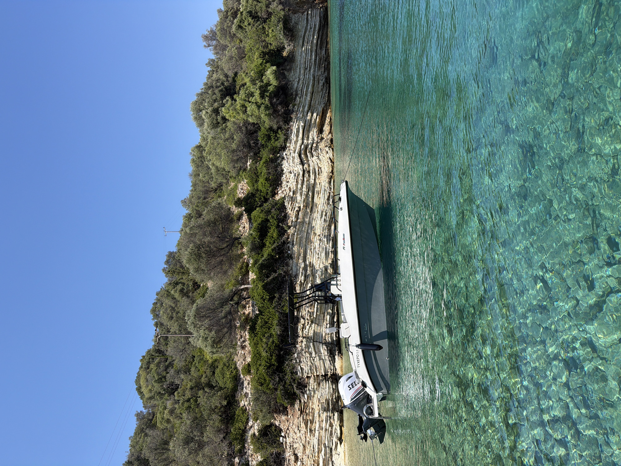 A small motorboat is moored in crystal-clear turquoise water off the coast of Kastos, Greece. The boat, a white and grey rigid inflatable boat (RIB) with a black canopy and outboard motor, is positioned in a cove with a rocky, layered cliff face and lush green vegetation covering the hillside. The water is so transparent that the rocky seabed is visible. The sky is a clear, bright blue. The scene depicts a serene and idyllic coastal environment.