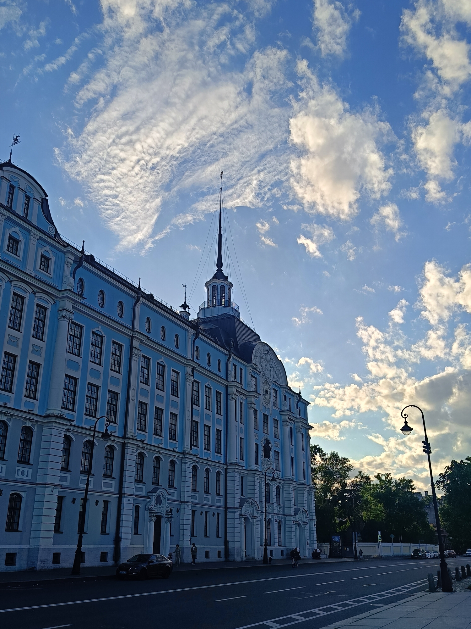 An exterior, eye-level, medium shot captures the side of a large, light-blue building with white trim and black-framed windows under a cloudy blue sky. The building, which appears to be historical, features multiple stories with arched and rectangular windows. A prominent spire with a weather vane tops a section of the building on the right side of the frame. A vintage-style streetlamp stands on the sidewalk in the foreground. A paved road with white lane markings runs alongside the building, with a parked black car and a few pedestrians visible. Trees and other structures are visible in the background.