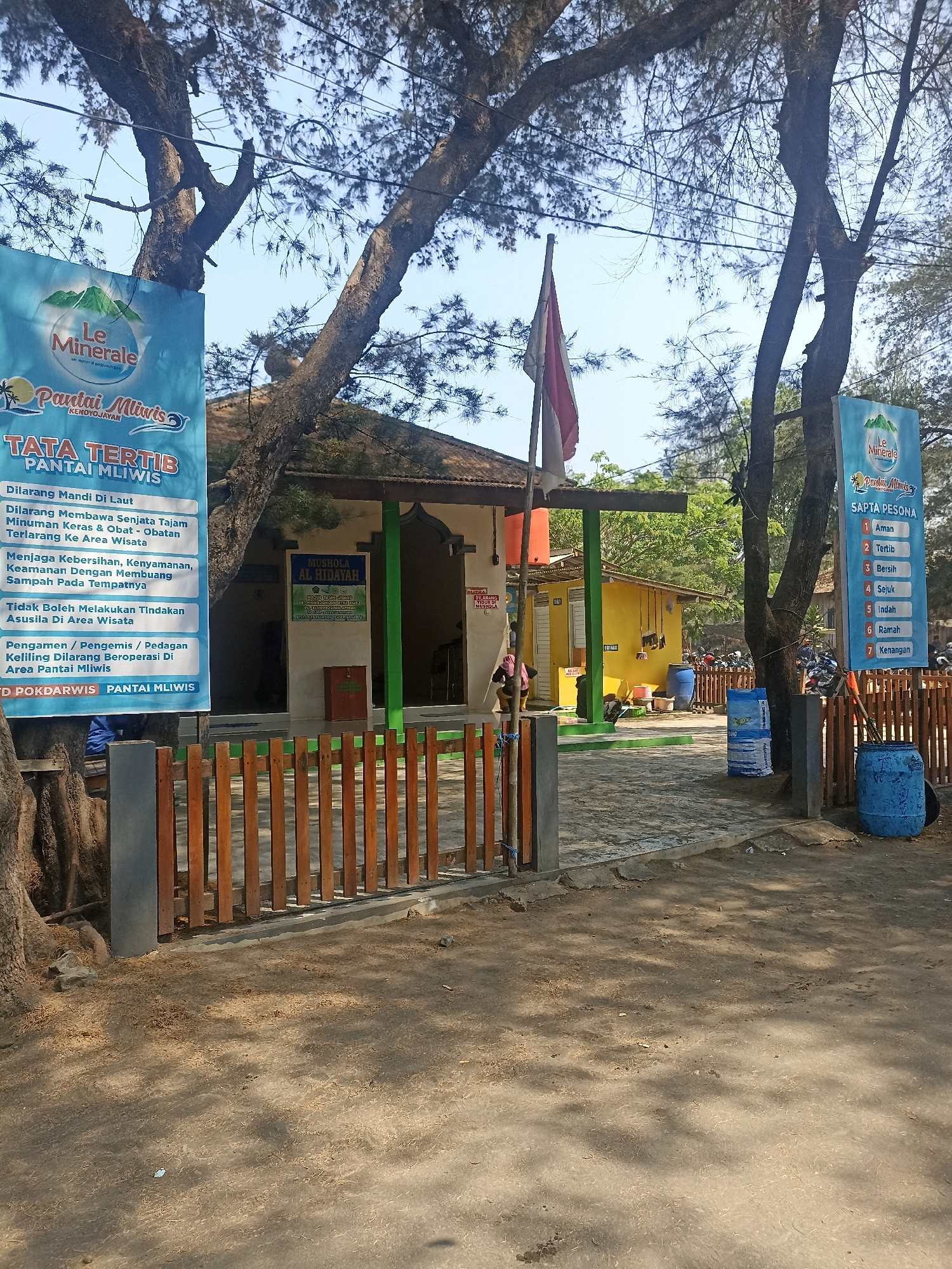 A vertical, eye-level, outdoor, full shot captures a beachside entrance in Kenoyajaya, Indonesia. The foreground is dominated by a sandy path shaded by trees, with a rustic wooden fence running along the right side. Two tall, blue plastic barrels stand on the right side of the path, one near the fence. The middle ground reveals a low-lying building with a yellow wall and a green-trimmed entrance, likely a facility. A red trash can is visible near the entrance. Above the entrance, a green sign reads "MUSHOLA AL HIDAYAH." To the left of the building, a blue banner with "Le Minerale" branding and beach imagery is attached to a tree. This banner displays "PANTAL MLIWIS" and "TATA TERTIB PANTAL MLIWIS," outlining rules for the beach. Further to the left, another identical blue banner is visible. A red and white Indonesian flag flutters from a flagpole in front of the building. The background shows more trees and a clear blue sky. Several motorcycles are parked on the paved area to the right of the building, suggesting visitors. A person wearing dark clothing and a pink head covering is partially visible near the building's entrance.