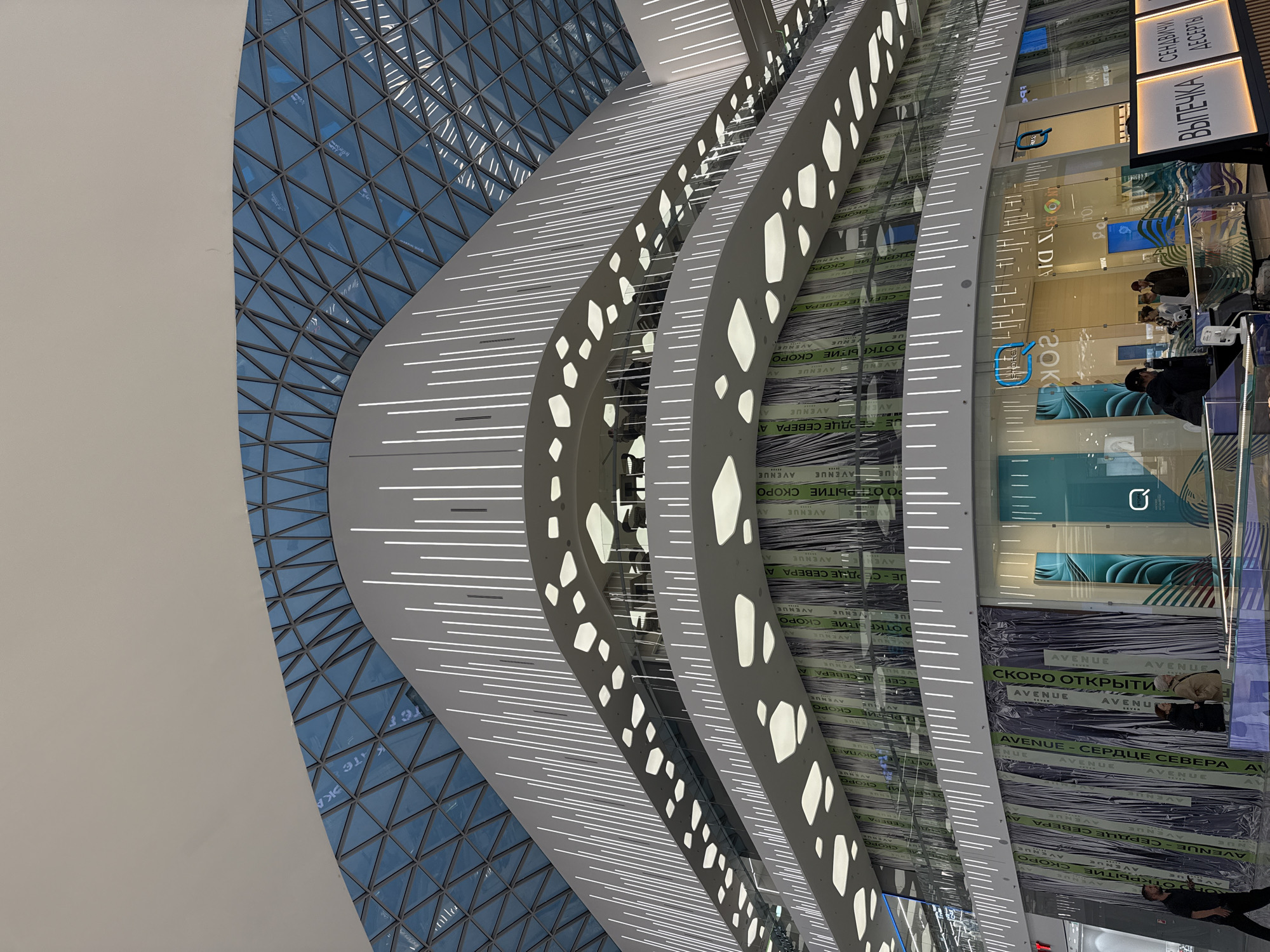 The image captures the interior of a shopping center, likely in Moscow, Russia, based on the Cyrillic script on the advertisements visible in the store windows. The architecture is modern, featuring a geometrically patterned glass ceiling and multi-tiered, curved walkways with illuminated linear designs and geometric cutouts. The walkways lead to storefronts, some of which are still under construction or preparing to open, as indicated by the banners that read "СКОРО ОТКРЫТИЕ" ("Opening Soon") and "AVENUE - СЕРДЦЕ СЕВЕРА" ("AVENUE - Heart of the North"). Several people can be seen inside the stores. Signage, "ВЫПЕЧКА СЕНДВИЧИ ДЕСЕРТЫ" ("Pastries Sandwiches Desserts"), indicate a food service establishment. The lighting is bright and contemporary, emphasizing the clean lines and modern design of the space.