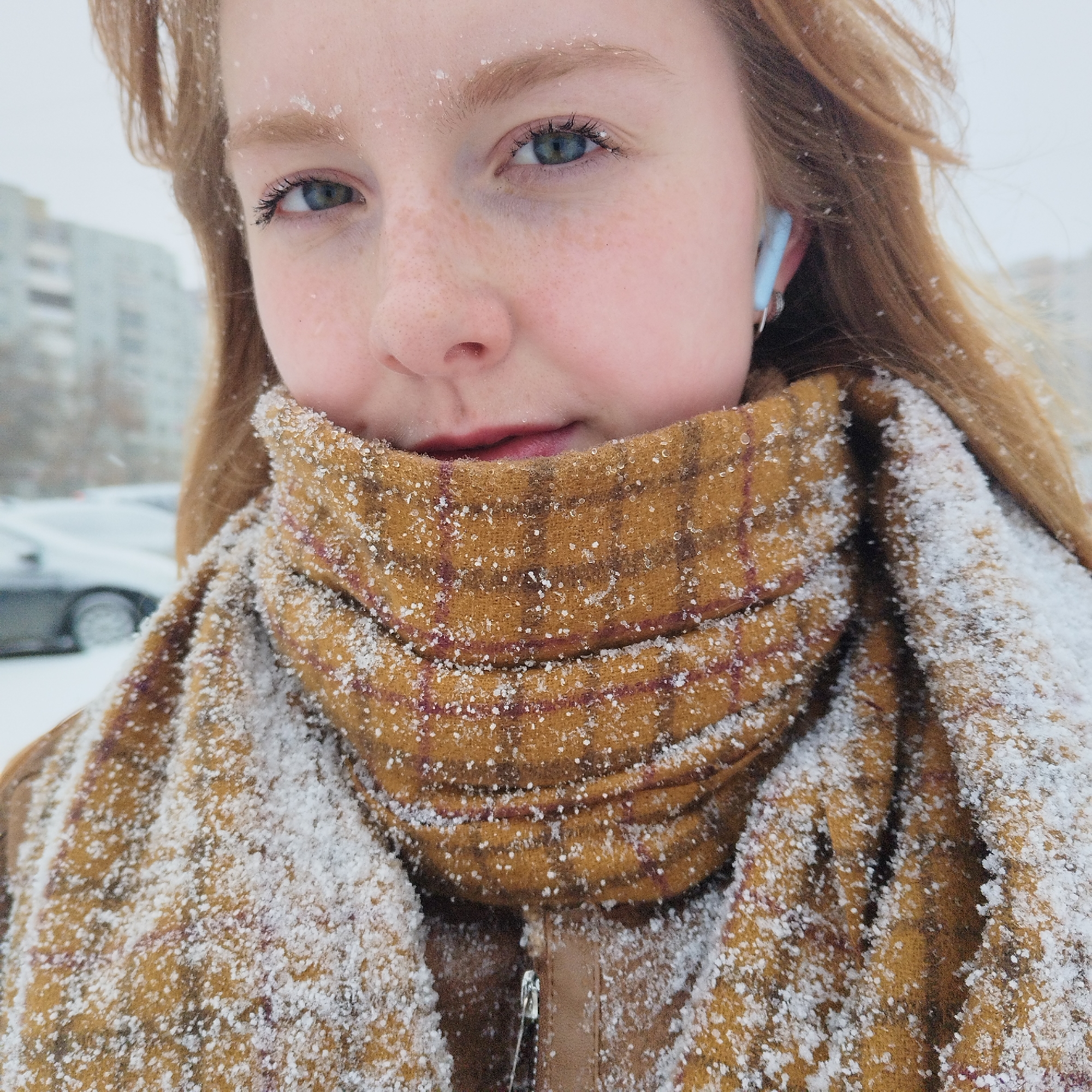 A young woman with red hair and light skin is centered in the frame, facing the viewer. She has blue eyes and freckles on her face. Snowflakes are visible on her eyebrows, forehead, and hair. She is wearing a brown and red plaid scarf wrapped around her neck and a brown jacket, partially visible. A light blue wireless earbud is in her right ear.
In the background, out-of-focus elements suggest a city setting in Yekaterinburg, Russia, with buildings and parked cars visible. The overall scene conveys a cold, snowy environment.