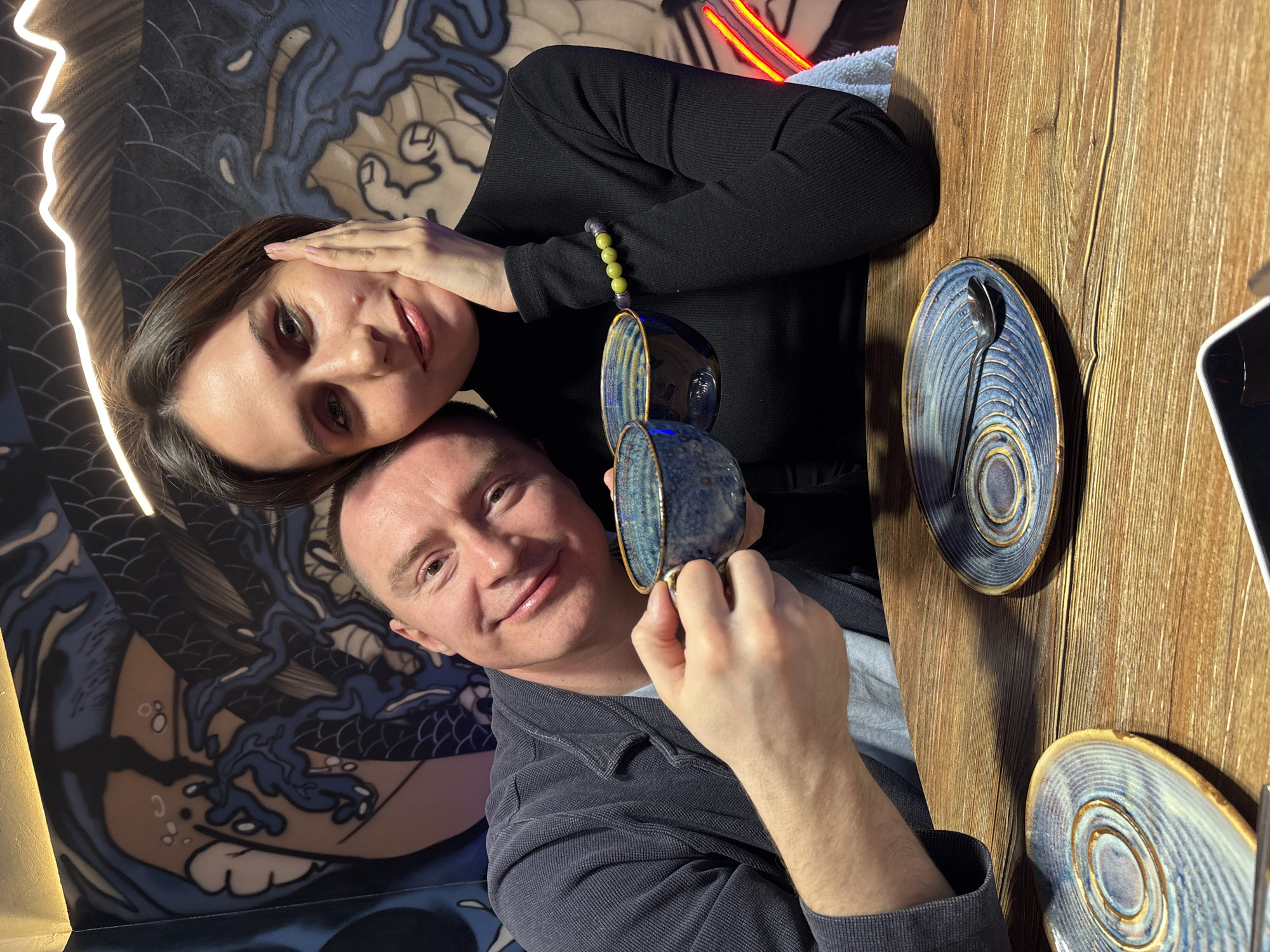 This image depicts two individuals, one male and one female, seated at a wooden table within an establishment in Izhevsk, Russia. The male, appearing to be in his 30s-40s with short brown hair, is wearing a light blue t-shirt beneath a dark grey textured cardigan. He is smiling and holds two dark blue, textured ceramic cups, one in each hand. The female, appearing to be in her 30s with short dark brown hair, is wearing a black ribbed turtleneck. She exhibits a faint smile, with her left hand resting against her left cheek. A beaded bracelet with green and purple spheres is visible on her right wrist. Both individuals have their heads leaning against each other, facing the camera. The table is light brown wood. Two matching dark blue ceramic saucers, each with a spiral pattern, are present on the table; one saucer contains a small silver spoon. The background wall features a mural with stylized blue and white wave or dragon motifs on a tan background. Decorative lighting includes warm strip lighting along the top and red neon-like elements further back. A white towel and a dark mobile phone are partially visible on the table's right side.