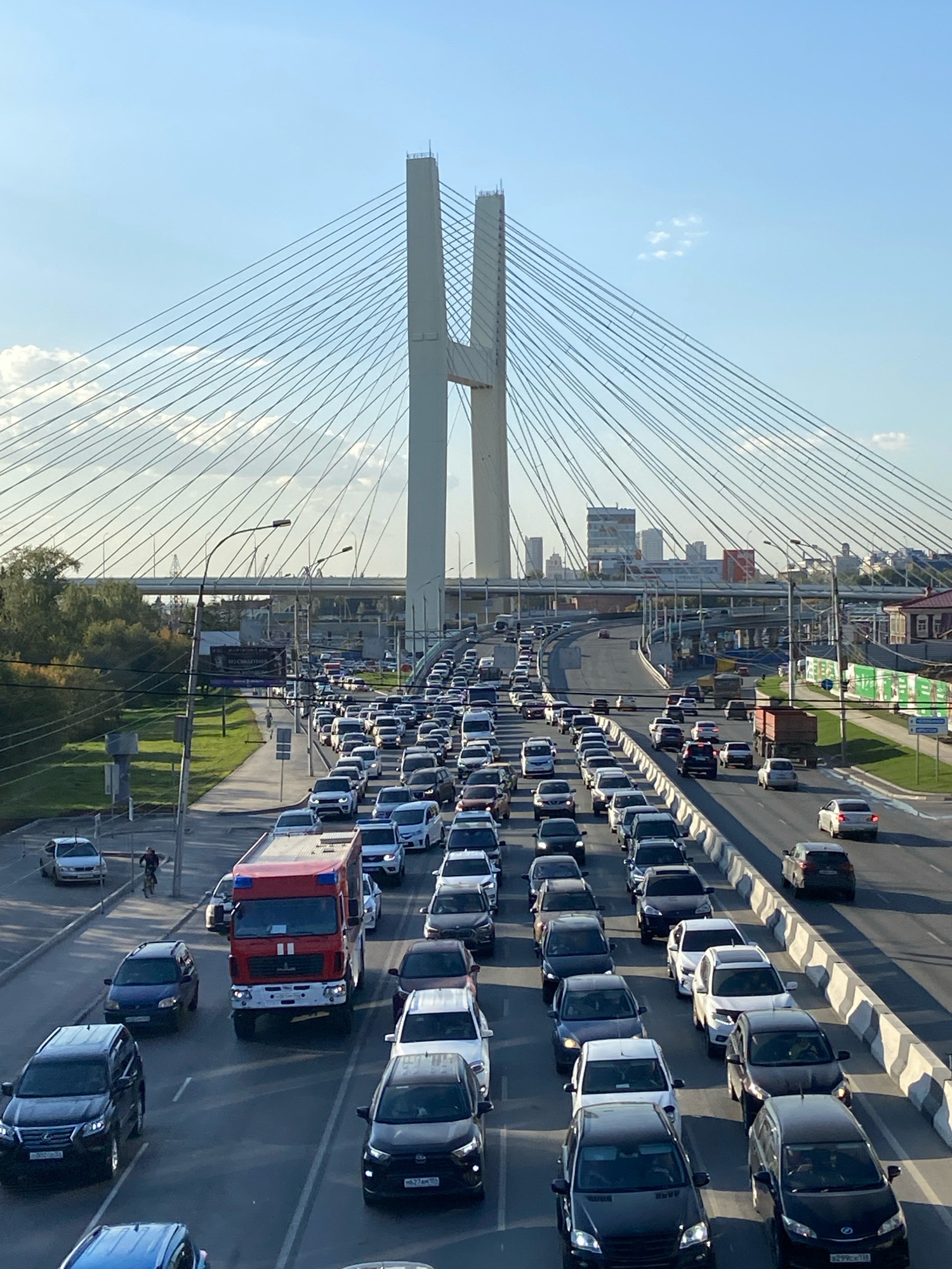 This is a high-angle, full shot of a congested cable-stayed bridge, identified as the Bugrinsky Bridge in Novosibirsk, Russia. The bridge spans a wide river, with its distinctive red arch structure visible in the background. The sky is a clear blue with scattered clouds. The bridge deck is filled with slow-moving traffic, forming long lines of cars, trucks, and a fire truck. The majority of vehicles are passenger cars, with a mix of colors and models. The roadway is divided into multiple lanes, separated by concrete barriers. On the left side of the bridge, there is a pedestrian walkway with a few people walking and cycling. Green trees and urban buildings are visible beyond the bridge. The scene captures a typical rush hour traffic situation on a major city artery.