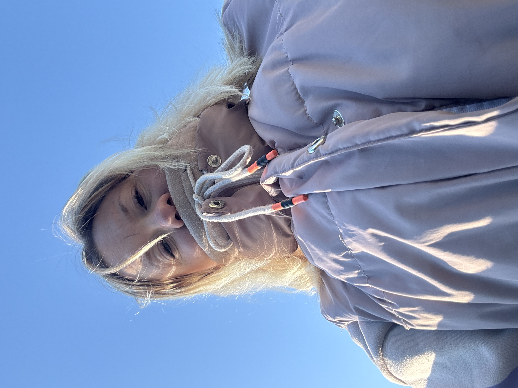 A woman with blonde hair, wearing a beige hooded coat, is photographed from a low angle against a clear blue sky. The hood of her coat is drawn up around her face with drawstrings. The location is outdoors in Ольховка, Russia.