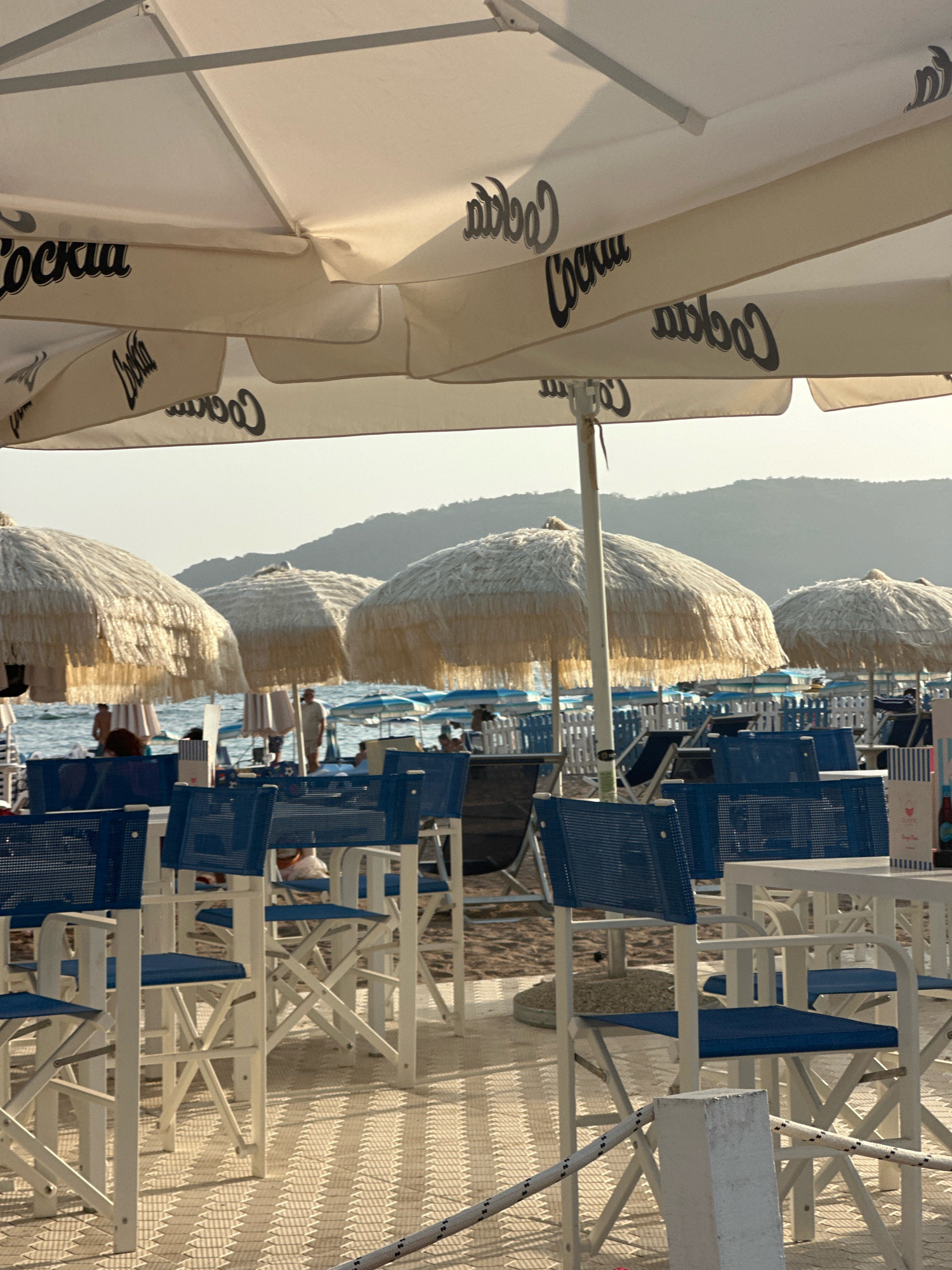 The image depicts a beachside cafe in Boreti, Montenegro, during what appears to be late afternoon or early evening, given the soft, warm lighting. The foreground is dominated by white tables and blue chairs, arranged on a patterned tiled surface. Several white umbrellas with black "Cockta" branding are open overhead, providing shade. In the mid-ground, more beach umbrellas, some with a thatched appearance, are set up on the sand. Beyond them, the calm sea is visible, with a few people walking along the shoreline and some beach chairs and umbrellas in the distance. The background is characterized by gently rolling hills that are bathed in the hazy sunlight. The scene conveys a relaxed and pleasant atmosphere, typical of a seaside resort.
