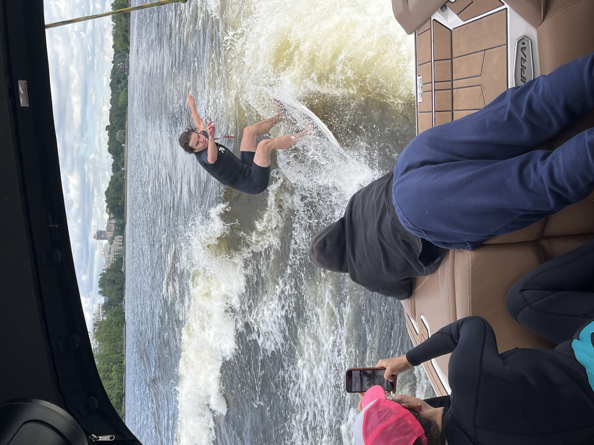 In this media file, a person is wakeboarding behind a boat on a body of water. The wakeboarder is wearing a black wetsuit and appears to be performing a trick, with their body twisted and arms extended. The boat, partially visible on the right, has brown upholstered seating and a section of teak decking. Two other individuals are seated on the boat, one in a dark hoodie and the other in blue pants. A person in the foreground is holding a smartphone, possibly filming or taking pictures of the wakeboarder. Another person in a pink hat is also visible. The scene is set on a river or lake, with a tree-lined shore and buildings visible in the distance, suggesting an urban or suburban environment. The weather appears overcast, with clouds filling the sky. The water is choppy, with a large wake generated by the boat. The overall impression is a recreational activity taking place on a somewhat cloudy day. The visible text on the boat reads "SUPRA."
