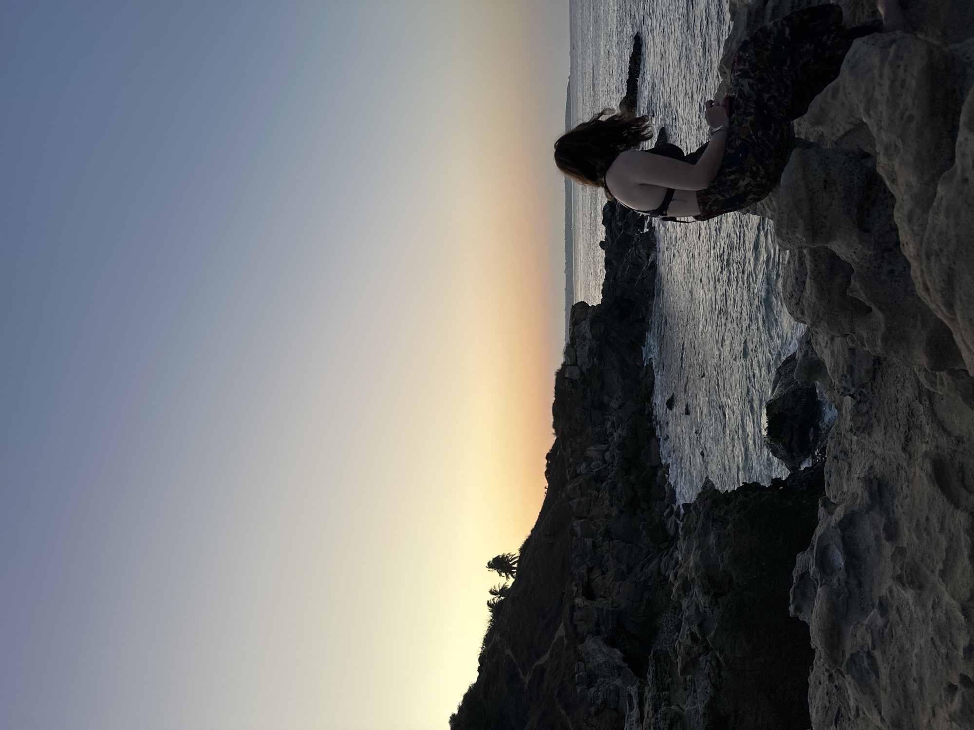 A woman is sitting on a rocky cliff overlooking the ocean at sunset in Puerto Escondido, Mexico. The sky is a gradient of light blue to yellow and orange where the sun is setting. The ocean is dark. The cliff is made of rough, light gray rock. The woman has brown hair and is wearing a dark top and patterned pants. She is holding a phone and looking at it.