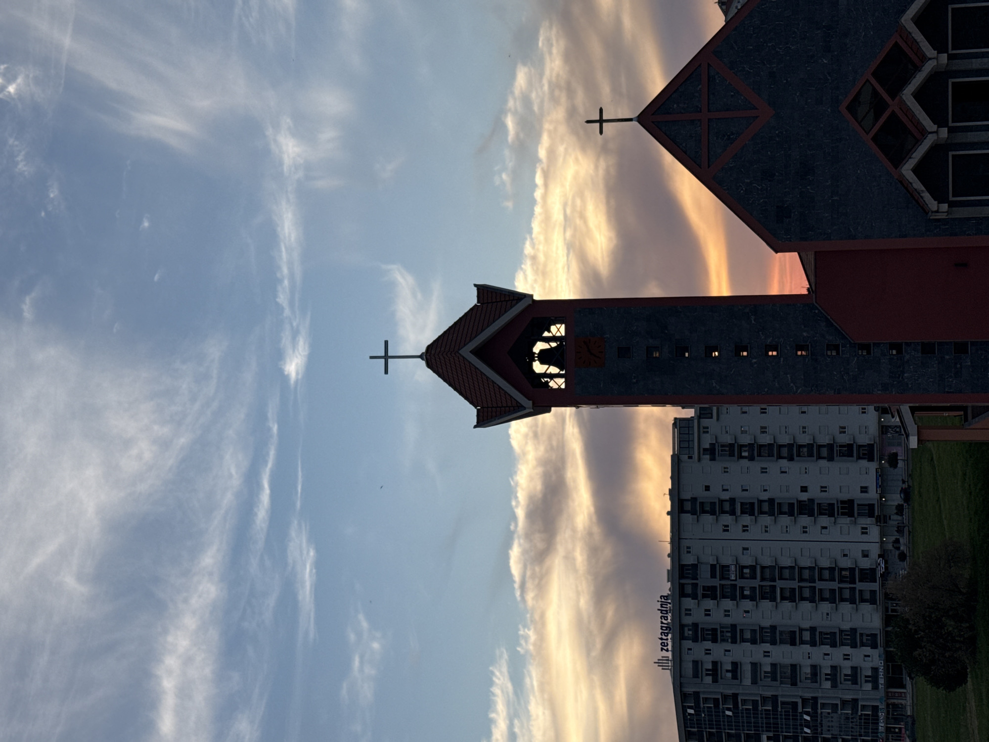 The image captures a low-angle view of the Church of St. John Vladimir in Bar, Montenegro, during what appears to be a sunset or sunrise. The church is characterized by its prominent bell tower and distinct architectural features, including its roof and cross. Adjacent to the church is a large, modern apartment building. The sky transitions from a deep blue to vibrant orange hues, indicating either dawn or dusk. Wispy cirrus clouds stretch across the sky. The image is rotated 90 degrees clockwise. The name "Zetagradnja" is visible on the apartment building.