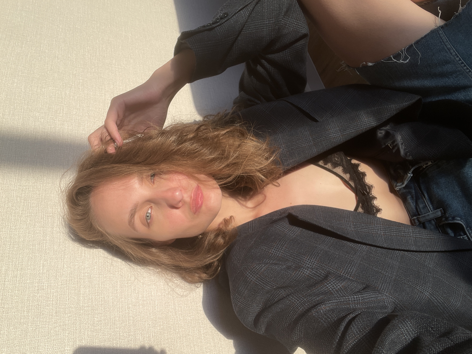 A woman with light brown hair and light blue eyes lies on a light-colored surface, wearing a dark, patterned blazer over a black, lace-trimmed garment and denim shorts. She is positioned at an angle, with her head near the left side of the frame and her body extending diagonally towards the upper right. Her left hand is slightly raised near her hair. The scene is lit by natural light, creating shadows. The location is unknown.