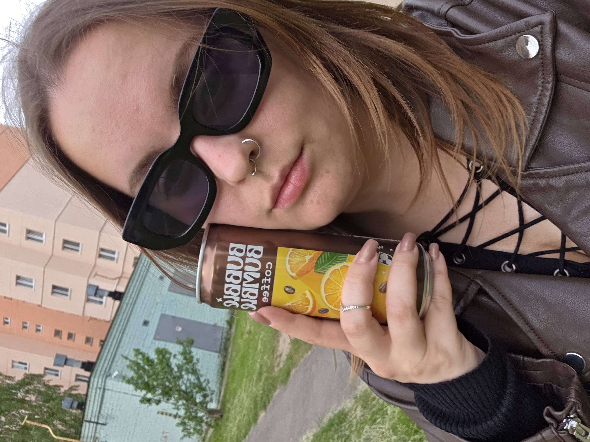 A female subject is depicted in a chest-up outdoor selfie, holding a can of "BUMBLE BUMBLE COFFEE" in her right hand. She has brown, shoulder-length hair and is wearing large black rectangular sunglasses that obscure her eyes. Her nose features a silver septum piercing consisting of one ring and one stud. Her lips are visible and appear light pink. She wears a brown faux leather jacket over a black top with visible criss-cross lacing on the chest. A silver ring with small stones is on her right ring finger, and her fingernails are painted a light brown/nude color. The can she holds is brown with a yellow and orange label displaying sliced oranges and coffee beans.
In the background, to the left, a light blue-green building with a dark roofline and ventilation structures is visible, with a small green tree in front. To the right, a taller, multi-story building with a light peach/pink facade and numerous rectangular windows is present. A paved pathway runs through a grassy area in the middle ground. The sky above is overcast. The setting is an urban or suburban outdoor environment.