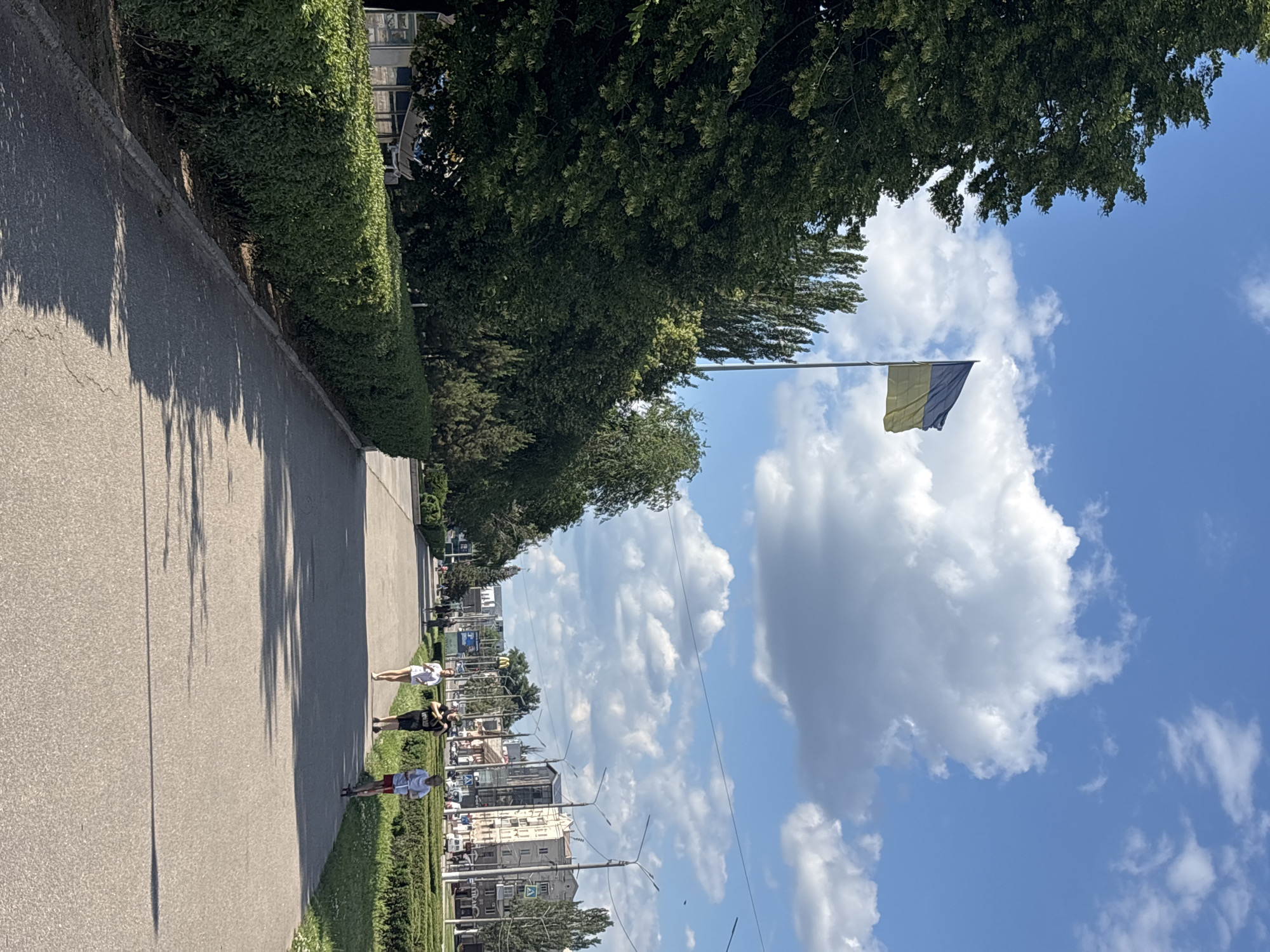 An eye-level, medium shot captures a sunlit day with a Ukrainian flag flying high against a partly cloudy blue sky. The flag is positioned on the left side of the frame, towards the top, and is gently waving. A pathway, paved with asphalt, stretches from the foreground into the distance, lined with neatly trimmed green bushes on the left and right. Several individuals are present on the pathway: two people are walking, one person is running, and one person is on a scooter. In the background, buildings of varying heights and architectural styles line the street, interspersed with trees and streetlights. A McDonald's sign is visible among the background structures. The overall scene depicts a public space, possibly a park or a wide boulevard, within an urban environment on a clear day.