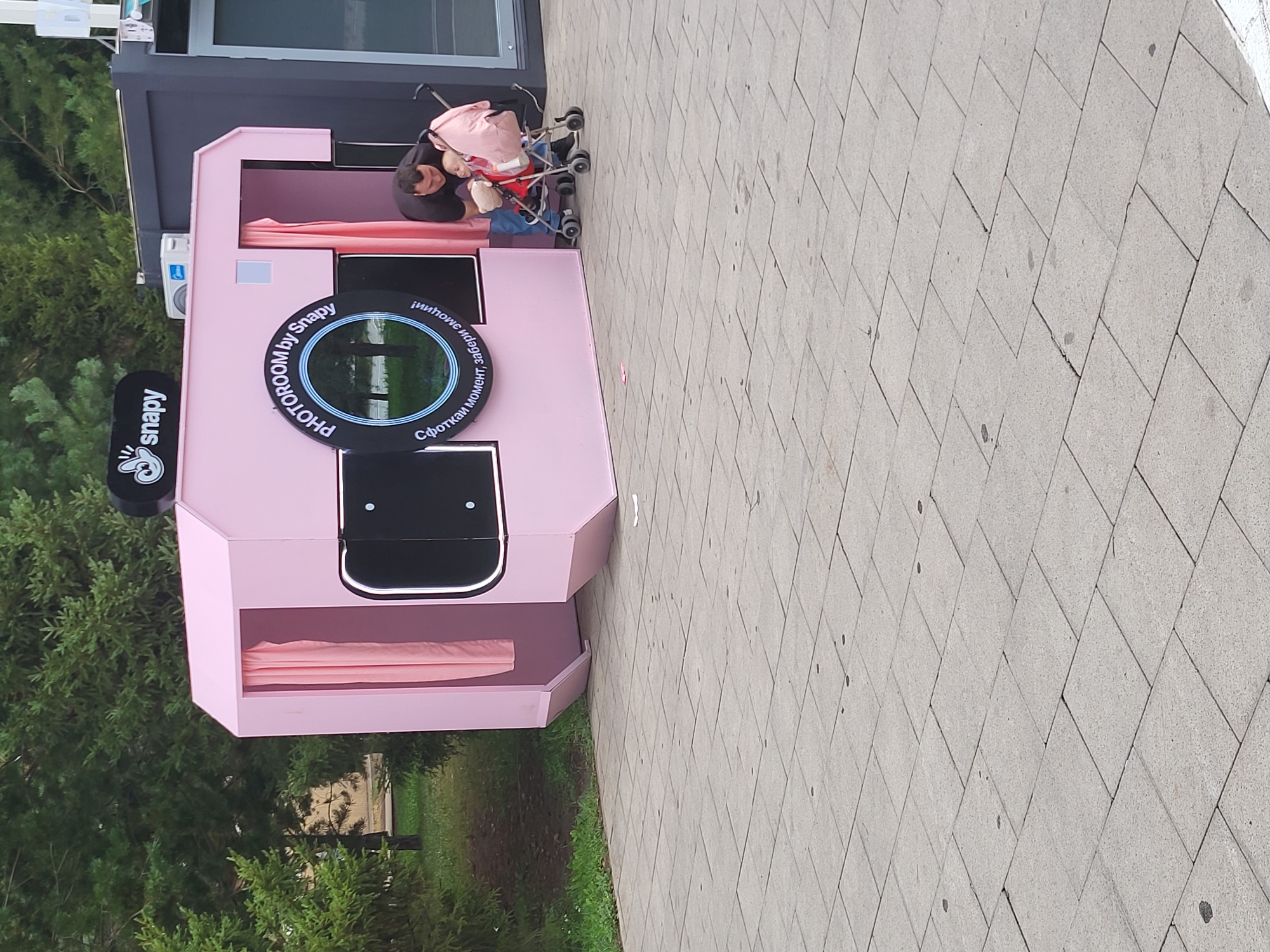 An outdoor, eye-level shot depicts a man bending over to interact with a baby in a stroller next to a pink photo booth. The photo booth is shaped like a camera, with a round opening showcasing a reflection of trees and sky. Text above the opening reads "PHỞTOROOM by Snapy". A logo with a cartoon mouse is visible on top of the booth. The man, wearing a dark t-shirt and jeans, is holding a baby who is in a pink stroller with a matching blanket. The stroller has four wheels and is positioned to the right of the photo booth. The scene is set on a grey tiled pavement, with trees and greenery in the background. The overall impression is a candid moment of a father and child near a recreational attraction.