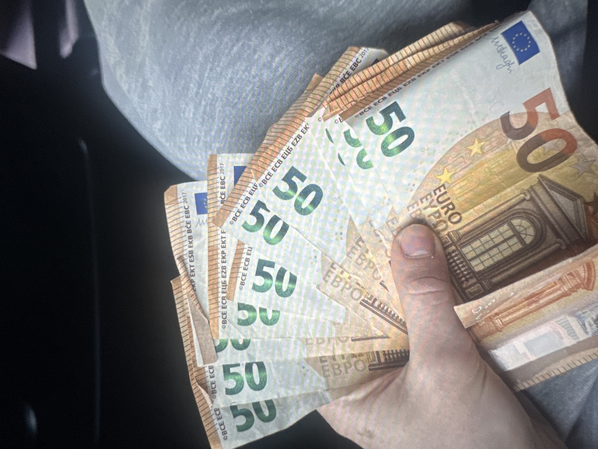 A hand holds a fan of several fifty euro banknotes. The banknotes are predominantly green and orange with visible text and serial numbers. The European Central Bank (ECB) and the year 2017 are visible on some of the bills, as are small stars and a watermark featuring a building. The background is dark and out of focus, with a hint of a light-colored textured surface. There is no indication of the location being in Tallinn, Estonia.