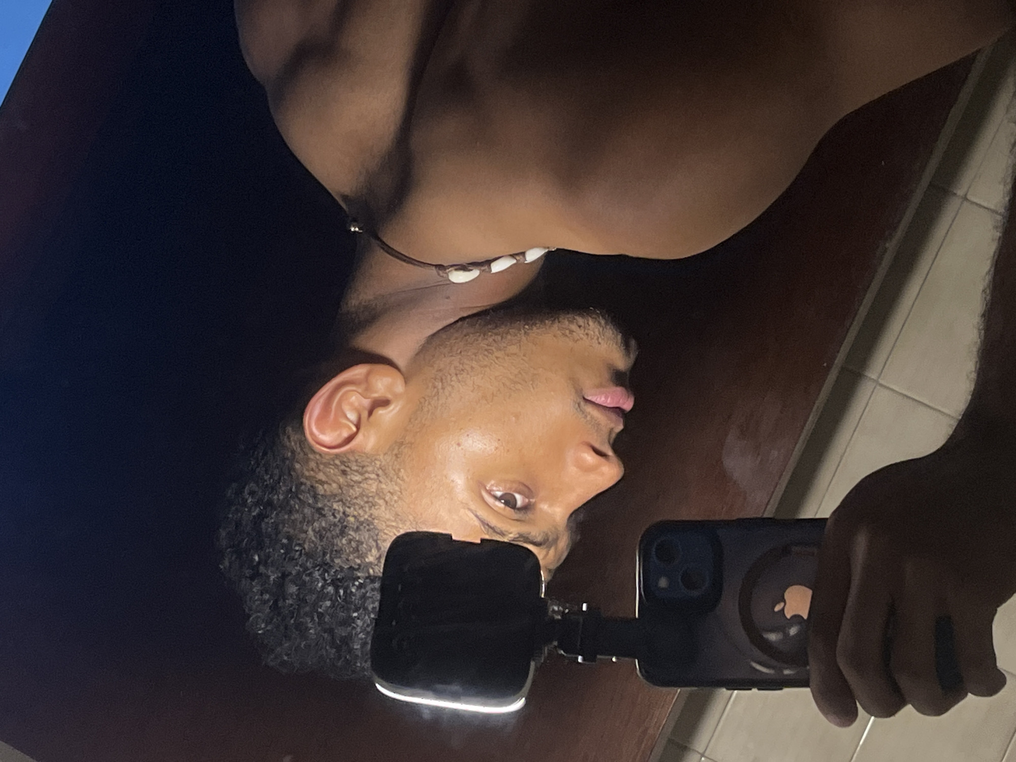 A tanned man is taking a selfie with an iPhone. The man is shirtless and is wearing a necklace. He has short, curly black hair and light brown eyes. The phone has a black case with an Apple logo. The background is dark. There is no indication that the image was taken in Porlamar, Venezuela.