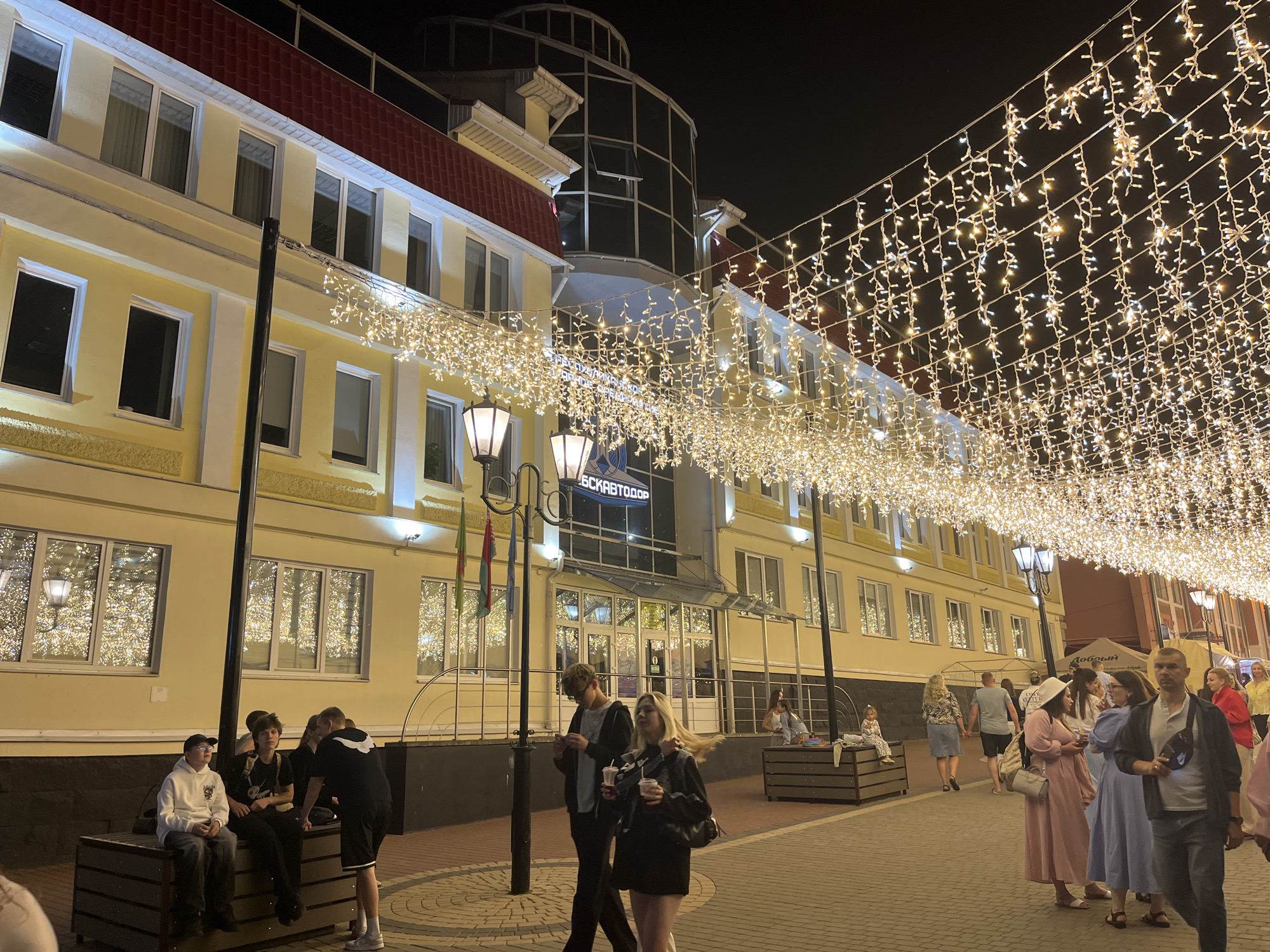 The image shows a street scene at night in Vitsebsk, Belarus, adorned with numerous strings of warm white fairy lights suspended across the street. A yellow building with a red roof dominates the left side of the frame, featuring multiple windows, some of which reflect the lights. Street lamps with traditional designs are visible. On the right side, more people are seen walking and standing, with some individuals in the foreground and middle ground. The ground is paved with cobblestones. A sign on the building reads "ВАСКАВТОДОР" (VASKAVTODOR). The overall ambiance suggests a festive or illuminated evening setting.