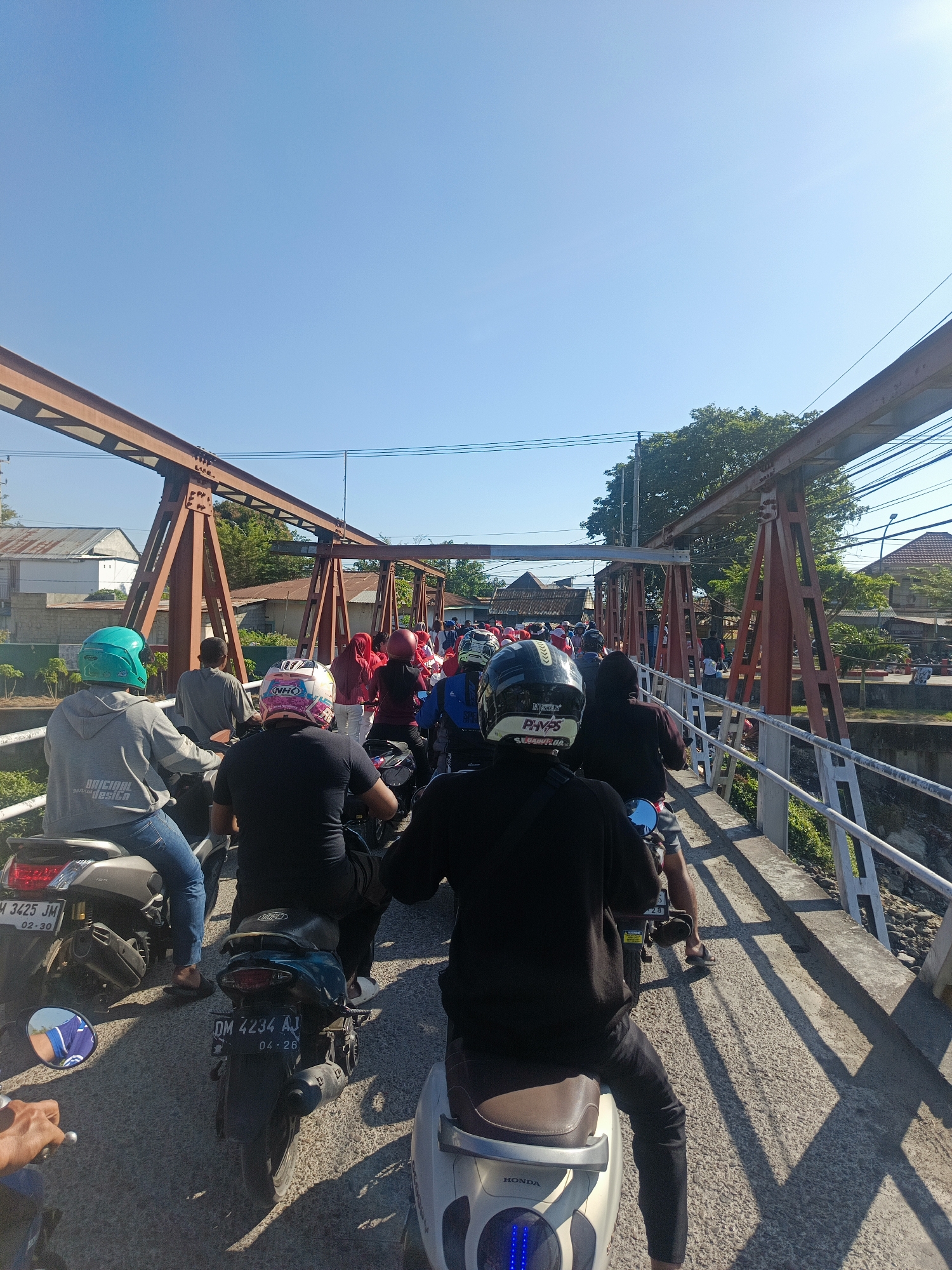 The image depicts a traffic jam on a bridge in Gorontalo, Indonesia. The bridge has a weathered, rust-colored steel truss structure. A line of motorcycles, with riders wearing helmets, is stalled on the bridge. The sky is clear and blue. Buildings and trees are visible in the background on either side of the bridge. The road surface appears to be asphalt. Some of the motorcycles have license plates visible, with one reading "DM 3425 JM" and another "DM 4234 AJ". The overall impression is a busy, everyday scene of congestion.