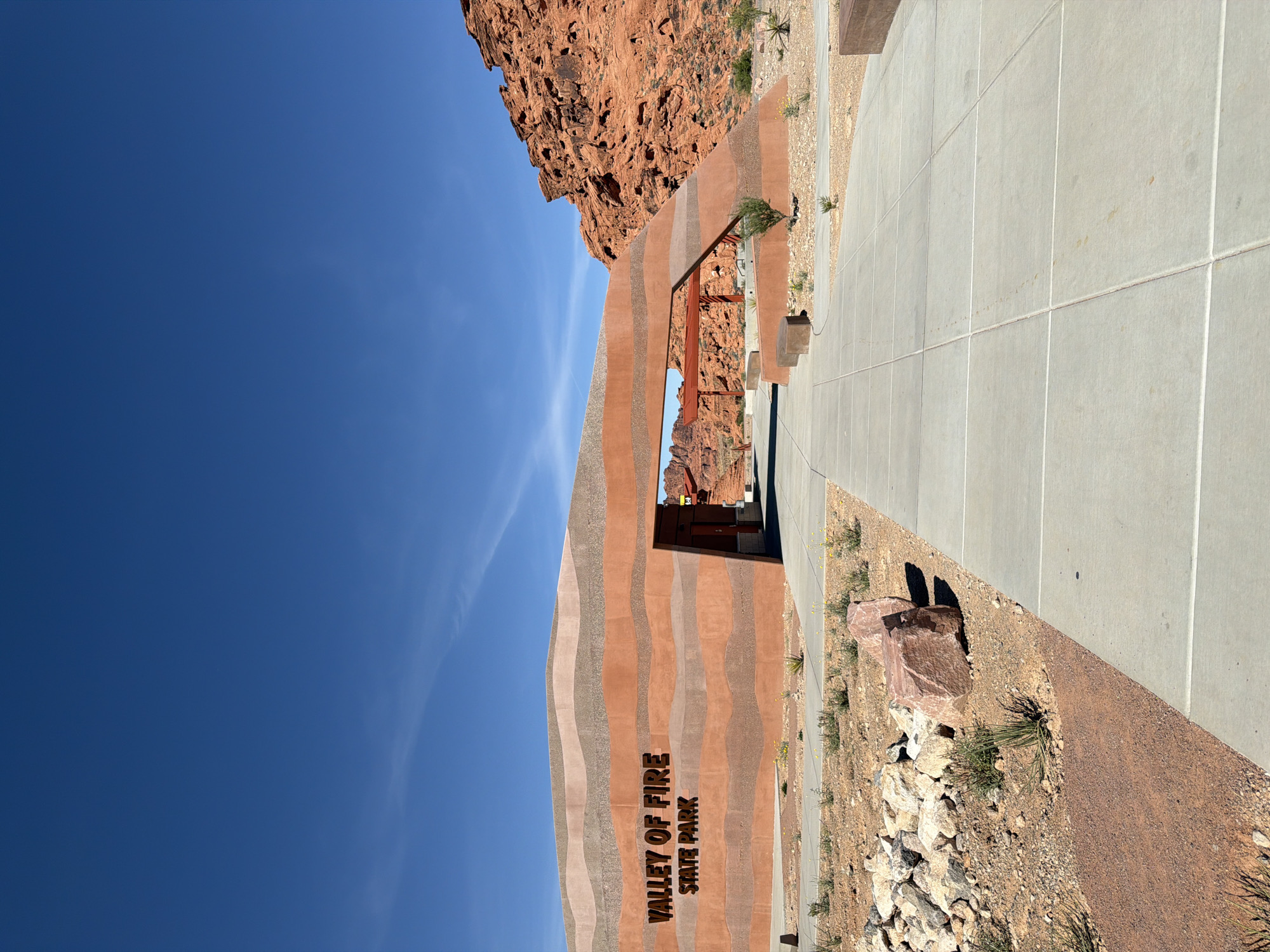 The image depicts the entrance to Valley of Fire State Park in Moapa Valley, Nevada, United States. The park's name is prominently displayed on the building, which has a striped pattern of alternating colors. There's a blue sky visible in the background. The building extends into the landscape on one side. There is a red rock outcrop on the other side, and a concrete sidewalk adjacent to the building. Small patches of desert vegetation are visible.