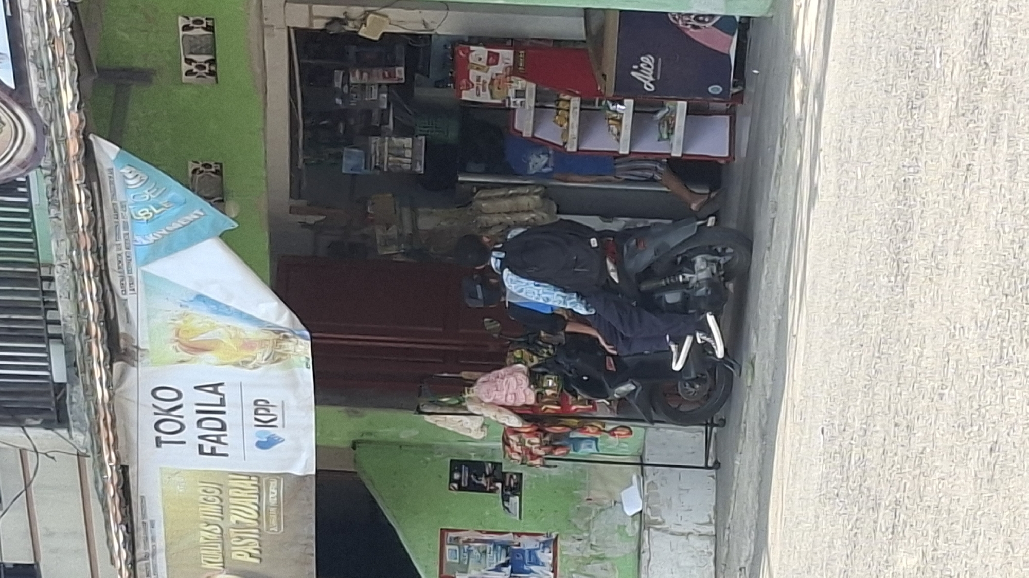 A street-level, medium shot captures a storefront in Cilaku, Indonesia, with a focus on two individuals on a motorcycle. The storefront, painted green, features a banner overhead displaying "TOKO FADILA" and "KPP" along with promotional imagery. Adjacent to the storefront, a red display rack holds various packaged goods, and a blue refrigerator unit is visible with branding that appears to say "Aice". Two people, possibly students due to their backpacks and attire, are seated on a black motorcycle parked in front of the shop. The person in front wears a dark cap and a blue patterned shirt, while the person behind has a black backpack and a blue and white patterned shirt. They are facing away from the camera, suggesting they are either leaving or arriving. The foreground shows a paved street with a shadow cast by the motorcycle. The background reveals more of the shop's interior, stocked with items, and a portion of a brown door. The overall scene depicts a typical commercial street in a residential area.