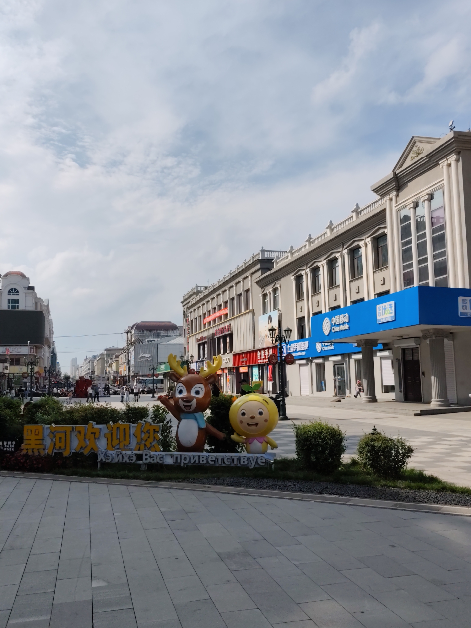 The image captures a street scene in Aihui, China, featuring a pedestrian walkway lined with buildings. In the foreground, a decorative sign with cartoon figures of a deer and a child welcomes visitors, with text in Chinese and Russian. The buildings on the right have a classical architectural style with columns and ornate facades, housing businesses like China Mobile. On the left, a mix of commercial buildings with varying architectural styles is visible, some displaying signage in Chinese. People are seen walking along the street and sidewalk, engaging in leisurely activity. The sky is partly cloudy, suggesting a bright day. The ground is paved with large gray tiles.