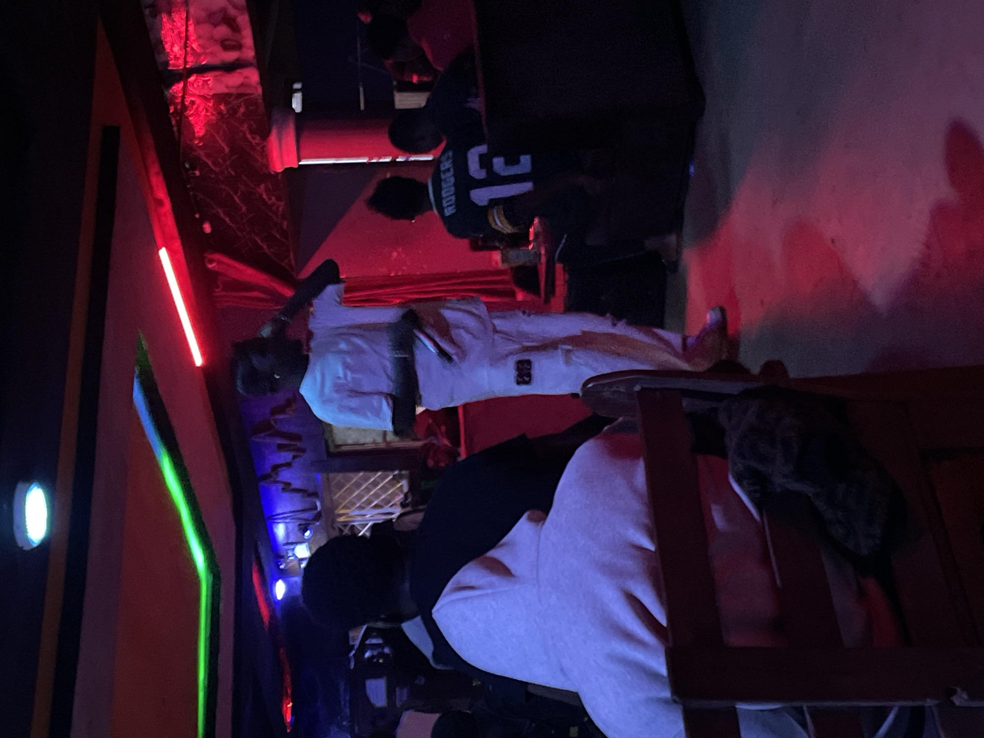 The image depicts an indoor entertainment venue, possibly a club or lounge, with distinct red and green neon lighting fixtures illuminating the ceiling and walls. In the foreground, a person is seated, facing away from the camera, wearing a light-colored hooded garment. To their right, another individual is visible, facing away and wearing a black shirt. In the mid-ground, a person in white attire is standing and appears to be speaking into a microphone, with their right hand near their mouth. This individual is also wearing white pants with a tag or patch on the right leg. Further to the right, another person is seated, wearing a dark green jersey with white lettering that reads "RODGERS 12". The floor is a light-colored concrete or polished surface reflecting the colored lighting. The background shows other individuals and furniture, including a dark-colored sofa. The overall ambiance suggests a lively, dimly lit environment. The specific location in the city of Durimi, Nigeria, cannot be definitively determined from the visual information alone.