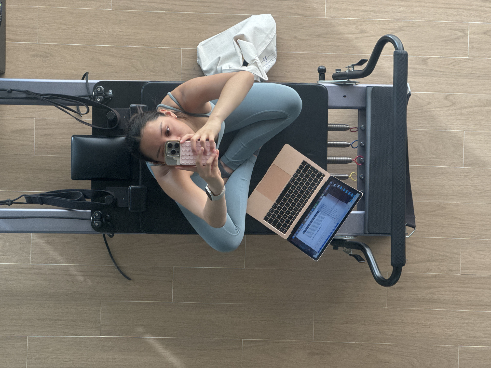 An overhead shot captures a young woman seated in a relaxed, cross-legged posture on a black Pilates reformer machine. She is taking a selfie with a silver iPhone, held by her left hand, which features a pink, textured case. A smartwatch is visible on her left wrist. She is dressed in light blue athletic wear, including a sports bra and leggings.
Next to her on the dark padded surface of the reformer, a rose gold or gold-colored MacBook Air laptop is open, its screen displaying text, likely from a document or webpage. Further to her left, a light-colored, possibly off-white canvas bag with a faint letter or symbol (resembling an "H") is placed.
The Pilates reformer is predominantly gray and black, with visible colored springs (red, blue, yellow) and black straps, indicating it's ready for use. The floor surrounding the reformer is covered with light brown, wood-look tiles, suggesting an indoor setting such as a home gym or a private studio. The lighting is bright and even, implying it is daytime. The scene depicts a moment of pause or multitasking during an exercise routine in San Bernardino Tlaxcalancingo, Mexico, with the woman appearing focused on capturing the self-portrait.