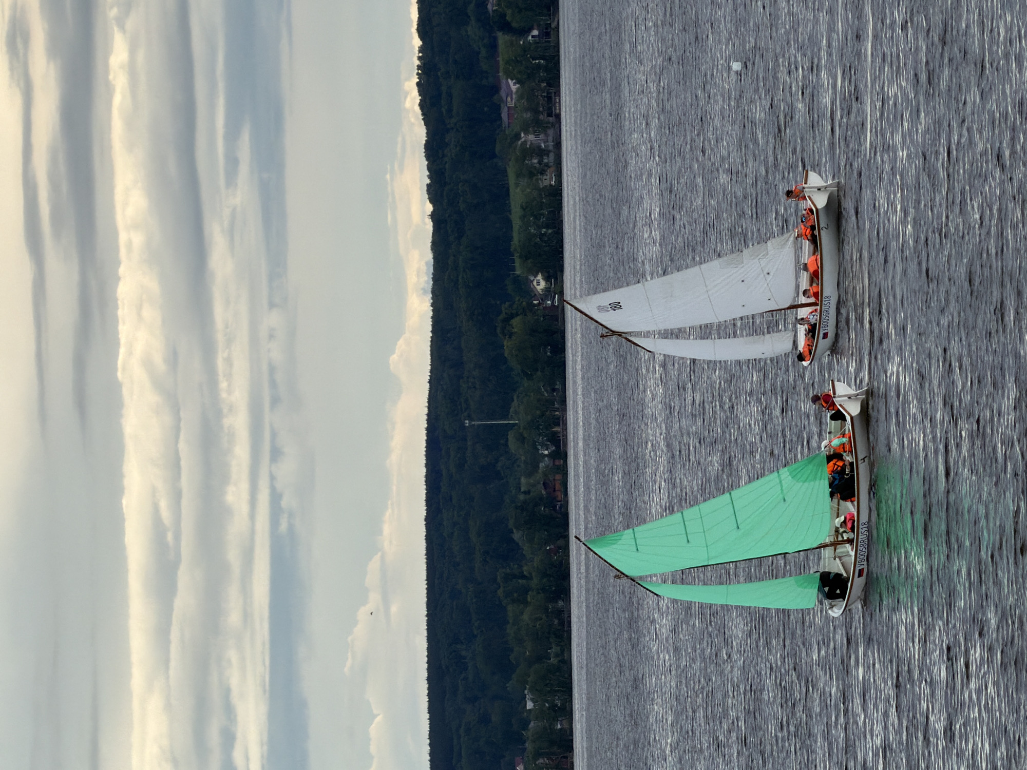 An aerial view captures two small sailboats navigating a calm body of water, likely a lake or reservoir in Votkinsk, Russia. Both boats are sailing away from the viewer, parallel to a distant, tree-covered shoreline.
The closer boat, positioned towards the lower right of the frame, features bright green main and jib sails that are fully unfurled. Several individuals wearing orange life jackets are visible within its white hull, actively engaged in sailing. On its stern, the text "V B0056RUS18" and the number "1" are visible.
Further away, the second boat displays white main and jib sails, also unfurled. It similarly carries multiple occupants, each wearing an orange life jacket. The number "068" or "088" is visible on its white main sail. Its stern also bears the text "V B0056RUS18" and the number "2".
The water's surface is a dark, rippled grey, reflecting the overcast sky and suggesting a light breeze. The background consists of a long, green hillside densely covered with trees, interspersed with several houses. The sky above is largely overcast with distinct, elongated cloud formations, indicating a late afternoon or early evening setting with diffused light. The scene portrays a peaceful recreational sailing activity.