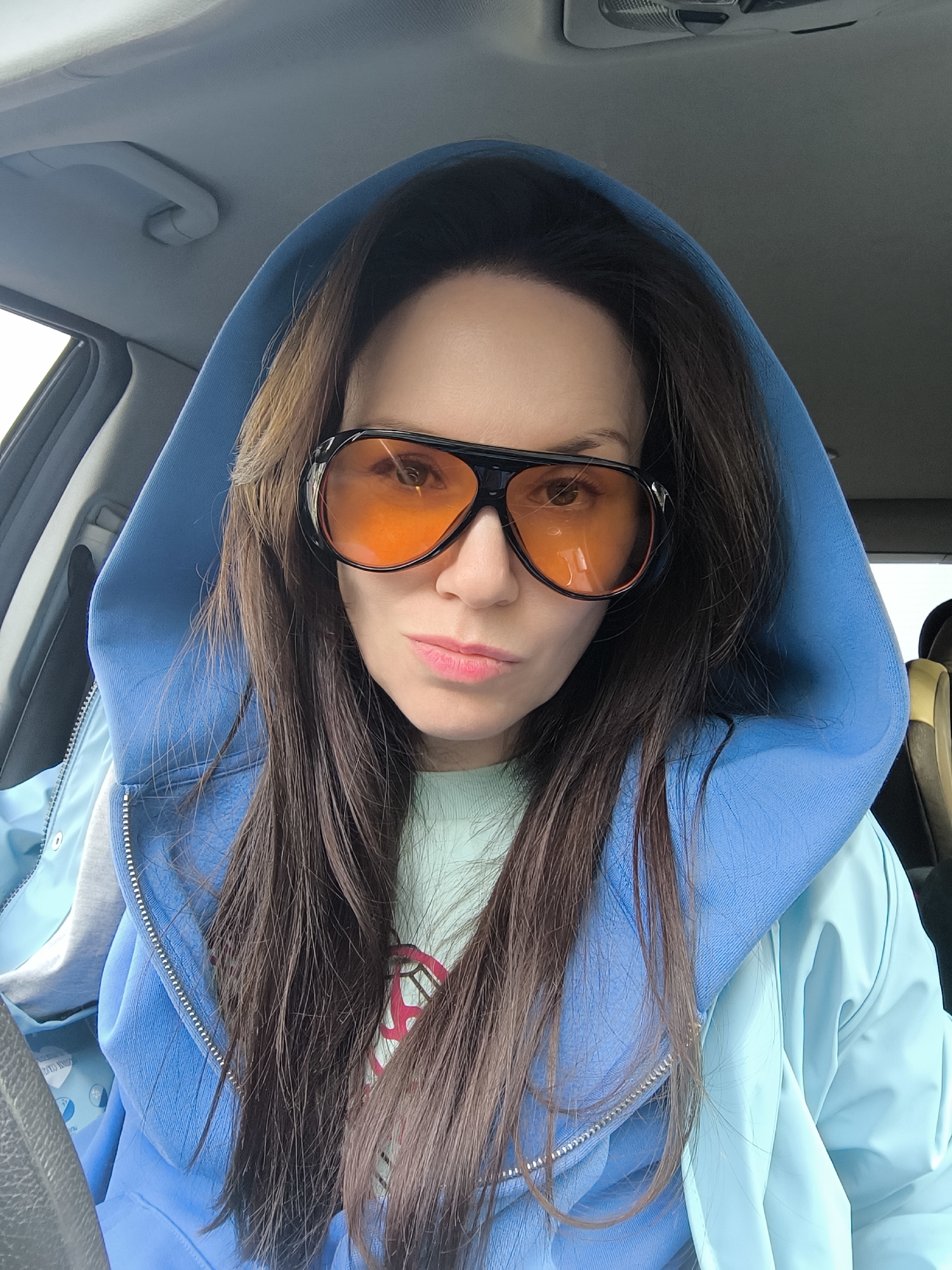 A woman with long brown hair is wearing a blue hooded jacket and orange-tinted glasses, taking a selfie inside a car. The interior of the car is visible, including the steering wheel and headrest of the driver's seat. The location is not evident, and there's no information suggesting this was taken in Riga, Latvia.