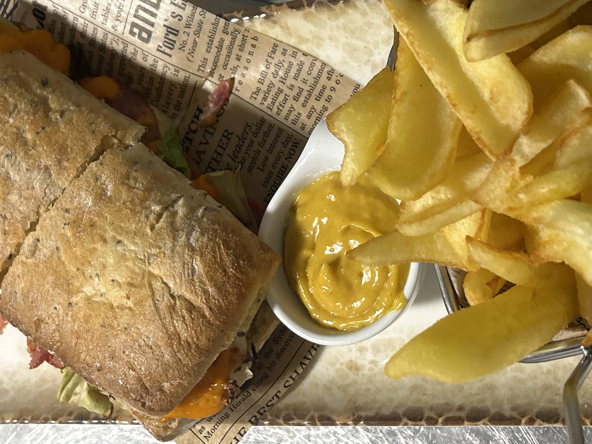 The image shows a close-up of a sandwich, fries, and a small dish of mustard on a tray covered with newspaper print. The sandwich appears to be made with a crusty bread roll and contains fillings like lettuce, cheese, and possibly meat. The fries are golden brown and piled high in a metal wire basket. The mustard is a smooth, yellow-orange color. The tray is lined with paper printed with newspaper-style text. There is no indication of a specific location in Burgas, Bulgaria, or any people present in the scene.