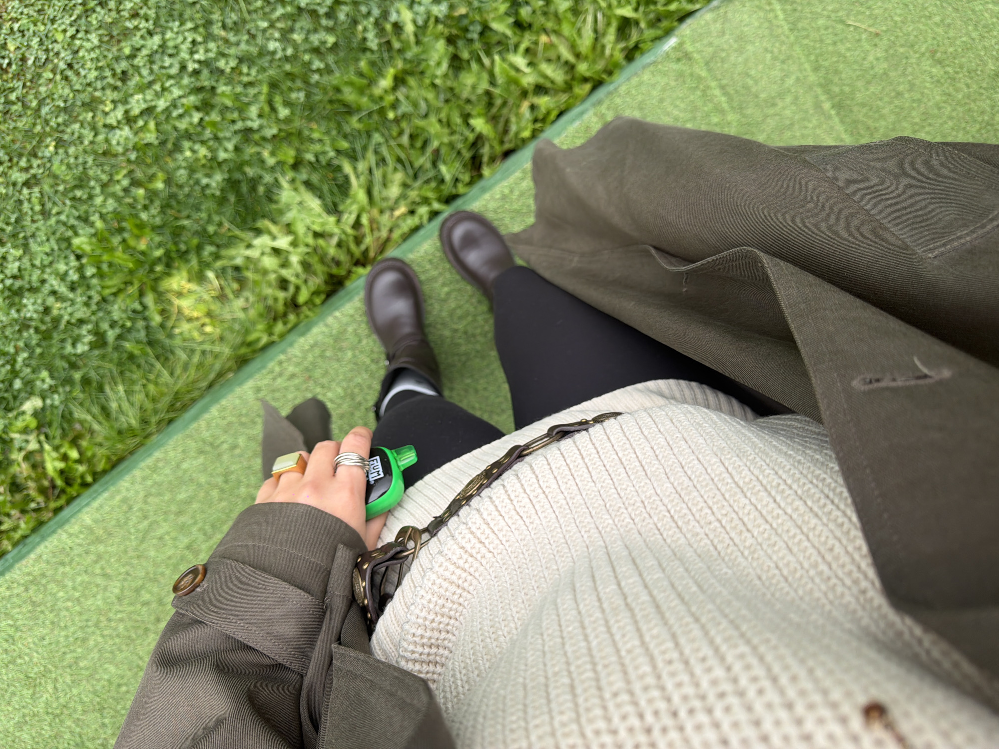The image shows a first-person perspective of a person sitting on a bright green surface, likely artificial turf, next to a patch of grass. The person is wearing a cream-colored knitted sweater, a dark green trench coat with a button on the cuff, black leggings, and dark brown boots. They are holding a small, bright green object, possibly a key fob or a small personal item, in their right hand, which also has several rings on their fingers and a large square-shaped ring on their index finger. A brown leather strap with metal detailing is visible across the sweater. The background consists of blurred green grass and foliage. There is no identifiable indication of the specific location within the city of Kamenka, Russia.