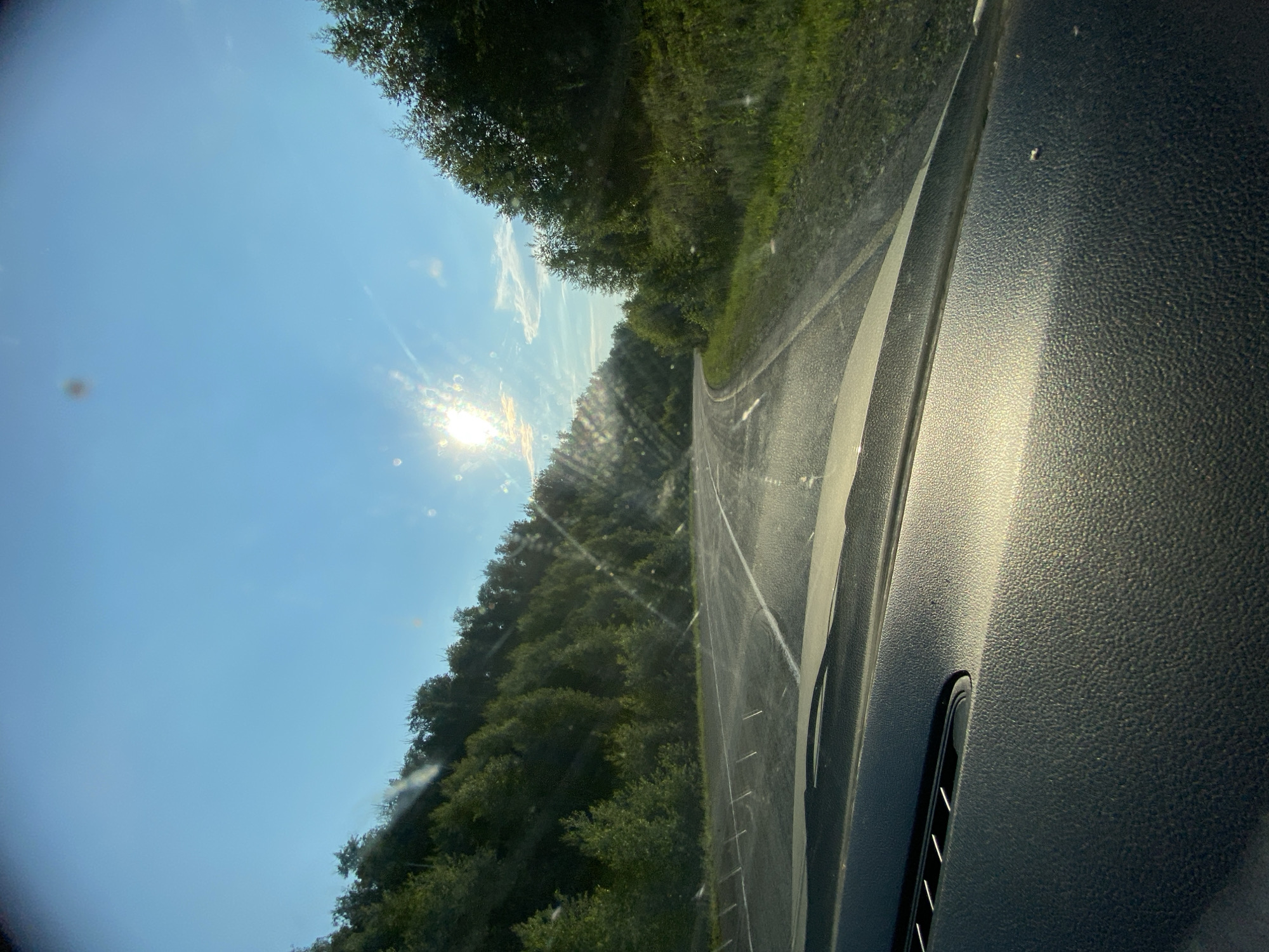 This media file captures a scenic view from inside a vehicle, likely on a rural road in Antingan, Russia. The image is oriented vertically, with the sun shining brightly in the upper left portion of the frame, casting sunbeams across the scene. The sky is a clear, bright blue with a few wispy clouds.
The foreground is dominated by the textured dashboard of the car, which is dark in color and reflects the sunlight. A portion of the windshield is visible, showing some smudges and lens flares, suggesting it might be slightly dirty or that the camera itself is producing these effects.
Beyond the windshield, a paved road curves to the right, bordered by a dense forest of green trees on the left and a grassy embankment on the right. The trees appear lush and full, indicating a warm season, likely summer. The road itself has white lines marking its lanes and appears to be in good condition.
The time of day is clearly indicated by the low angle of the sun and the warm, golden light illuminating the dashboard and the edge of the road, suggesting it is either early morning or late afternoon. The weather appears to be sunny and clear. There are no people or explicit text visible in the image. The overall mood conveyed is one of a peaceful, sun-drenched drive through nature.