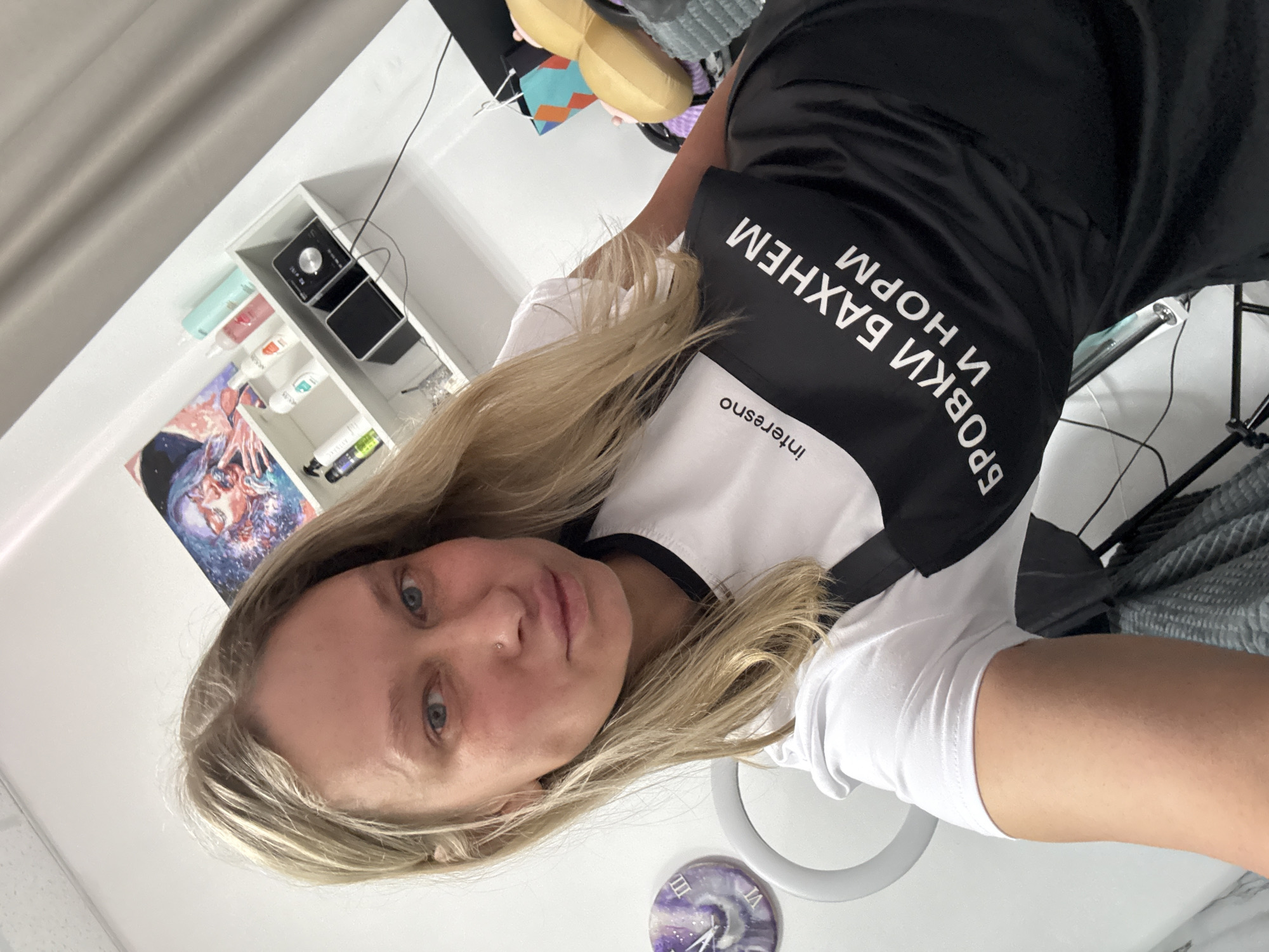 A waist-up selfie shows a blonde-haired, fair-skinned woman in Perm, Russia, wearing a white T-shirt and black apron. The apron has the text "interesno" and the phrase "БРОВКИ БАХНЕМ И НОРМ". Behind the woman are a white shelf unit holding items and a framed piece of art. There is also a ring light and a decorative clock on the table. In the background, there appears to be a light-colored wall and miscellaneous objects.